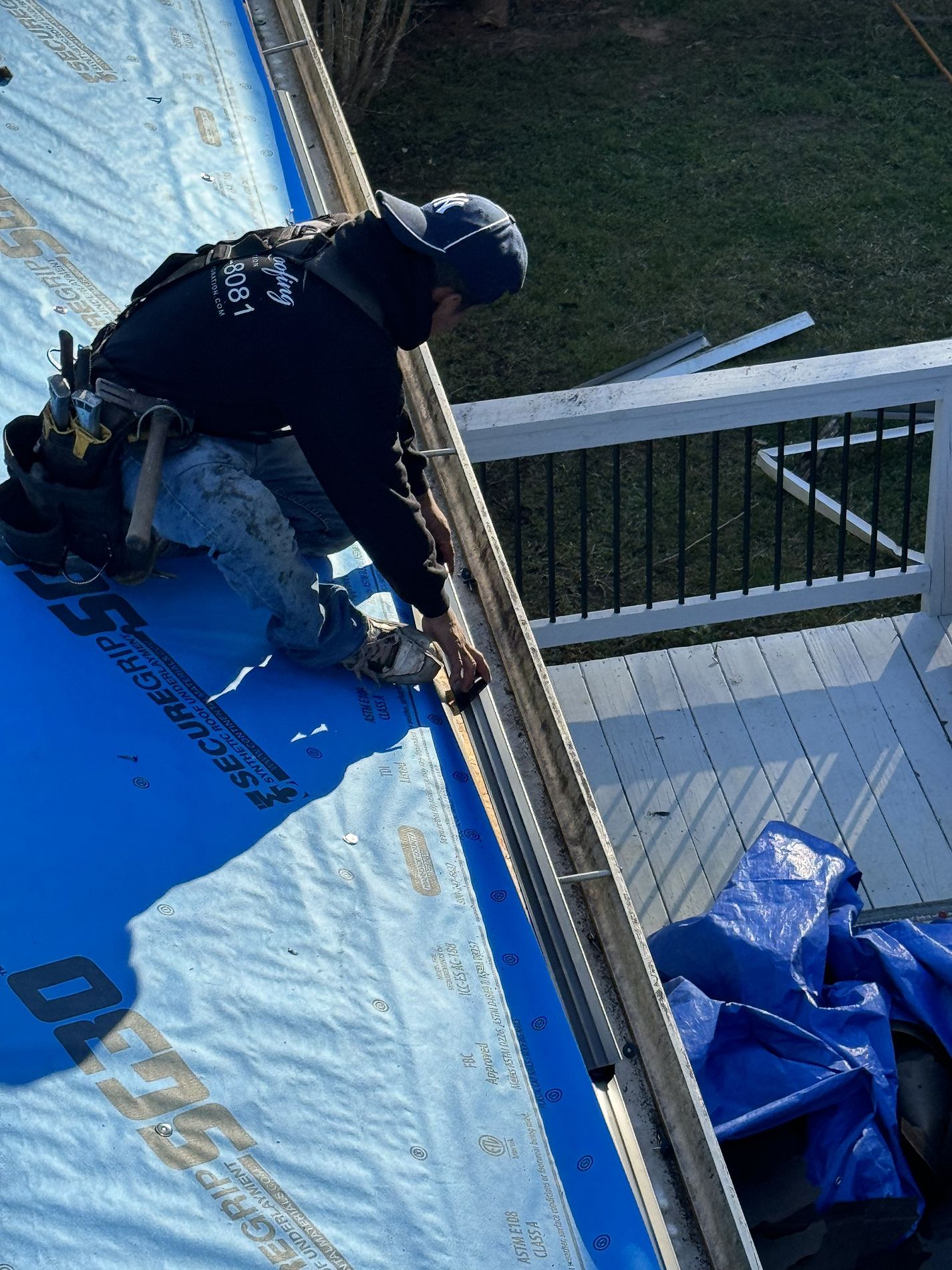 Man wearing a cap and tool belt working on roof gutter installation over a blue waterproof underlayment.