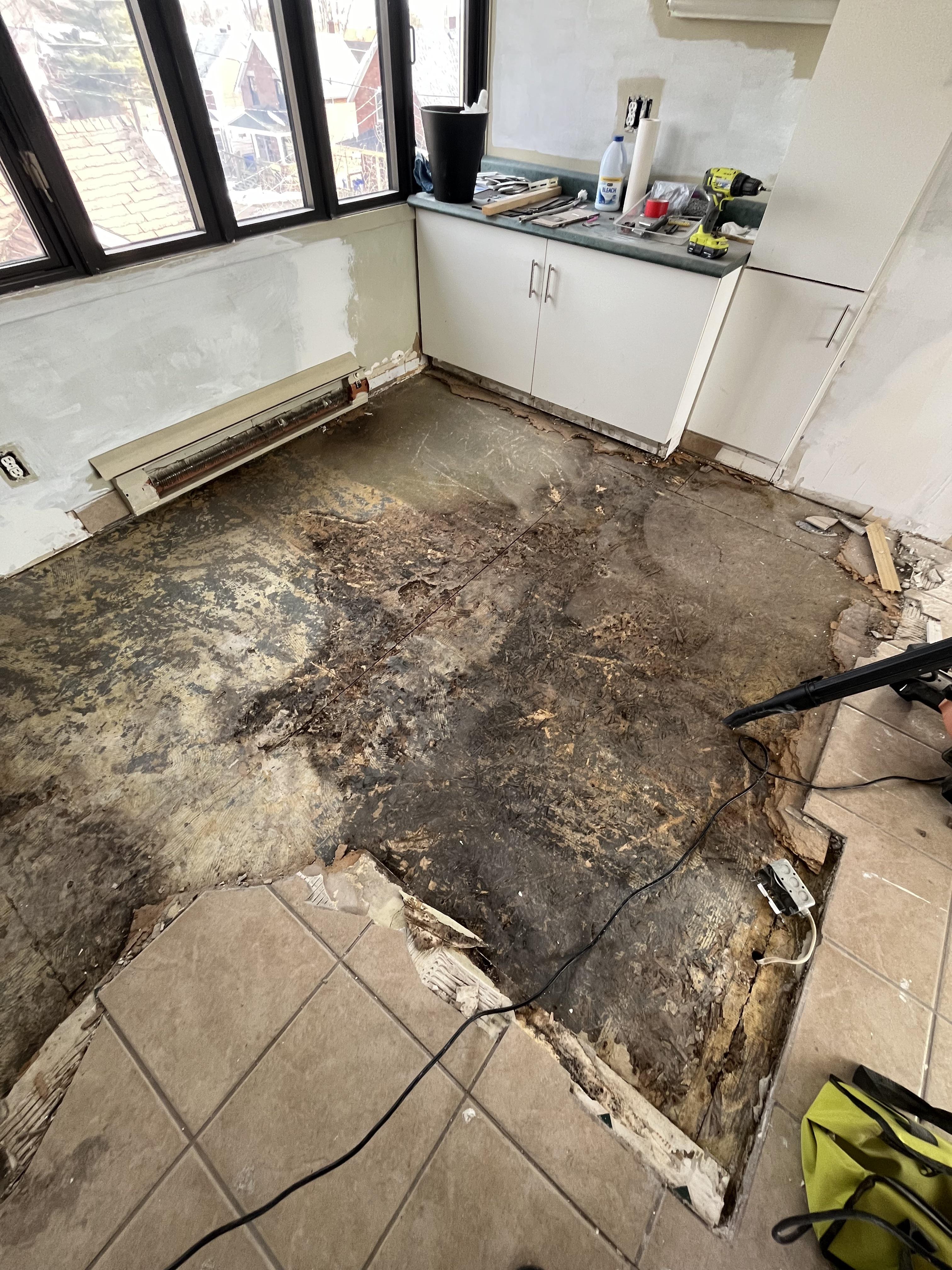Partially removed floor tiles revealing damaged, darkened subfloor with exposed wiring near kitchen cabinets.