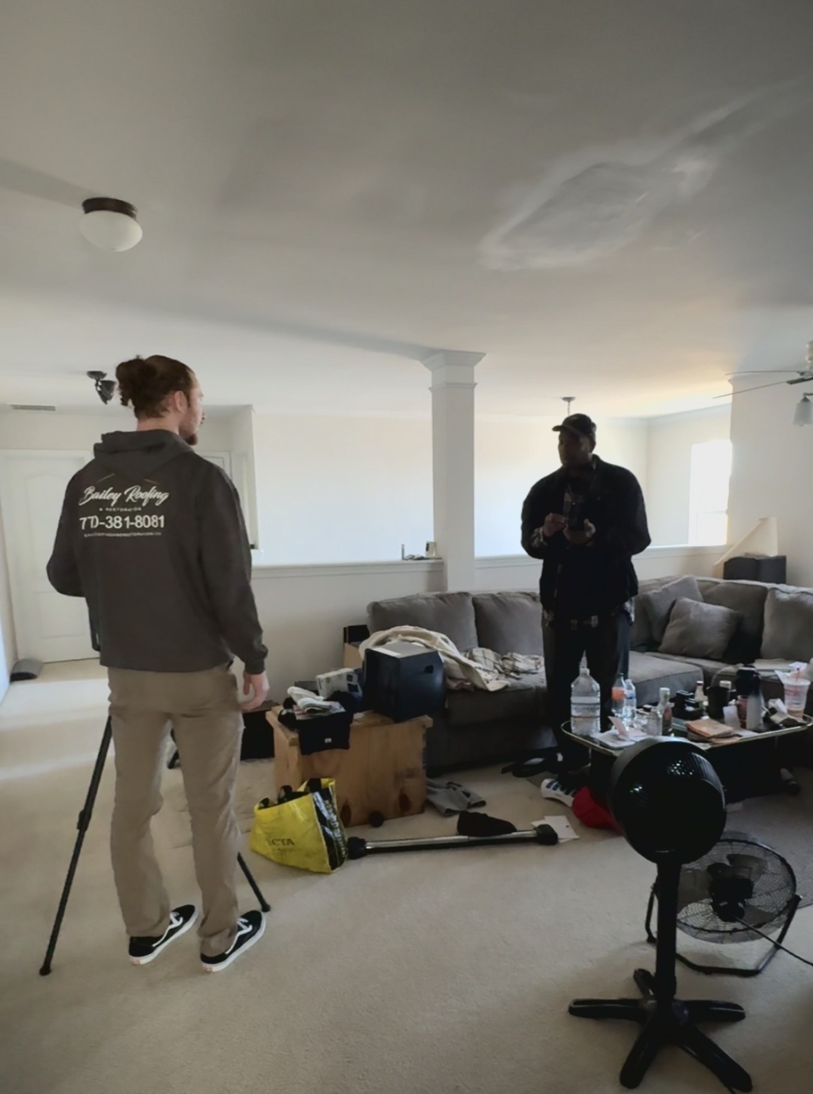 Two men standing and talking in a living room with a gray sectional sofa, cluttered coffee table, standing fan, and various items on the floor.