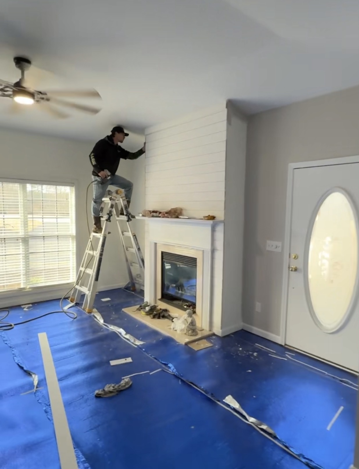 Man on a ladder installing white wooden panels above a fireplace in a living room with blue protective floor covering.