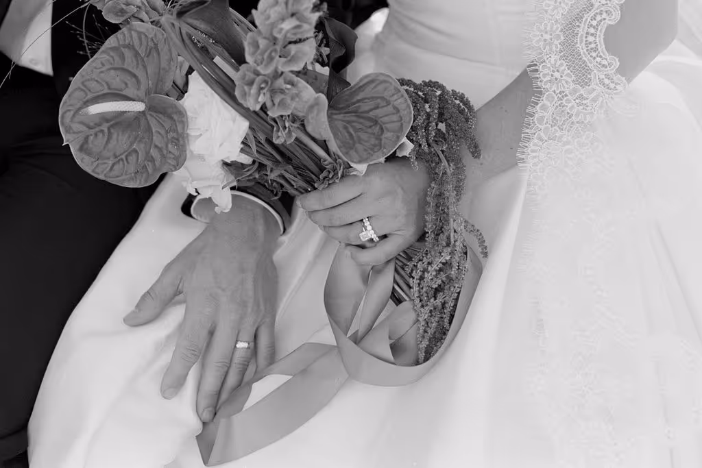 Close-up of a bride and groom's hands resting on the bride's white wedding dress, showing their wedding rings and holding a bouquet with ribbon.