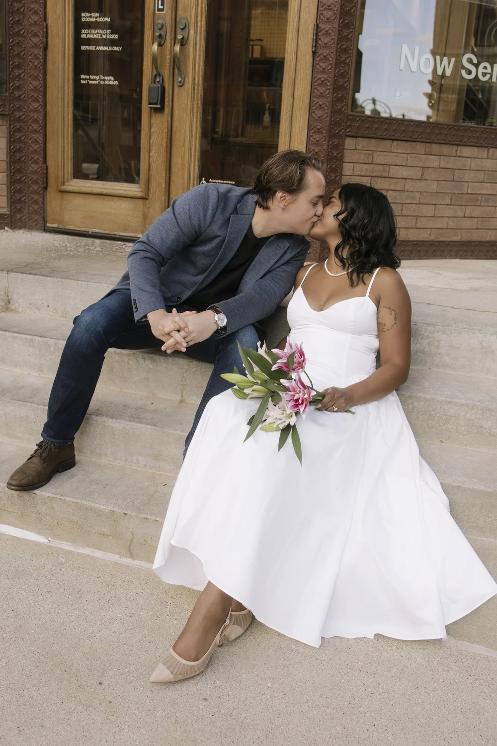Couple sitting on outdoor steps kissing, woman in white dress holding pink lilies bouquet.