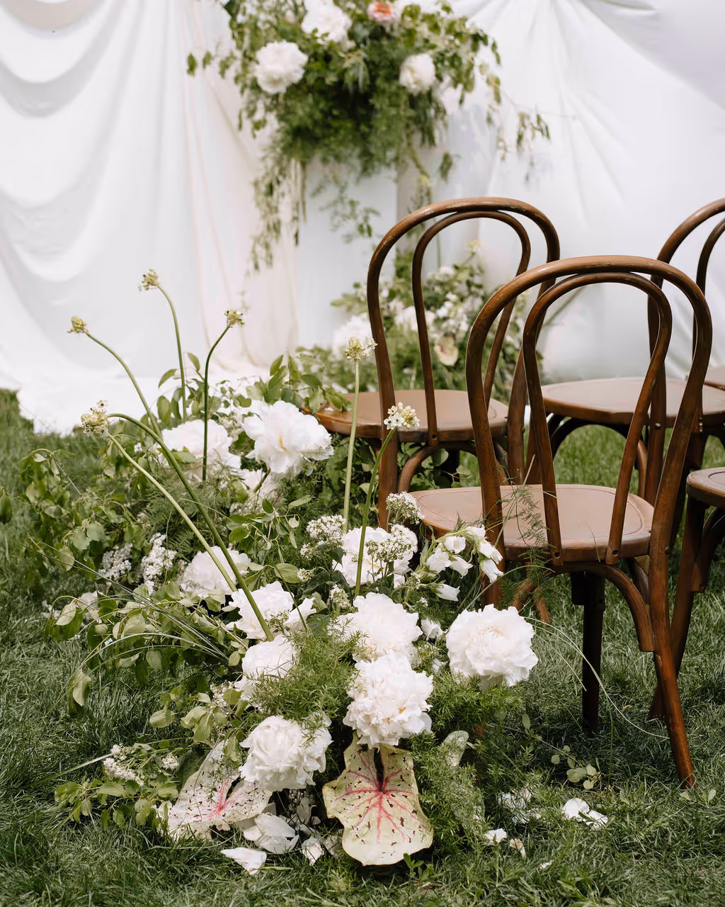 Elegant outdoor wedding setup with wooden chairs and lush white floral arrangements on green grass.