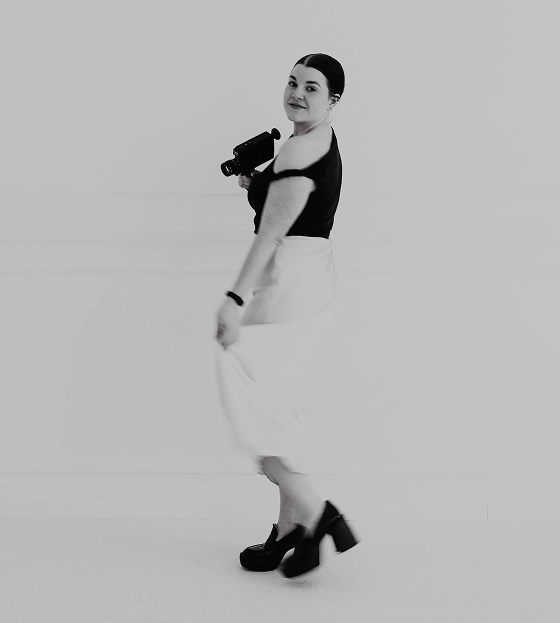 Laura in black top and white skirt holding a camera, looking back and smiling in a minimalist setting.