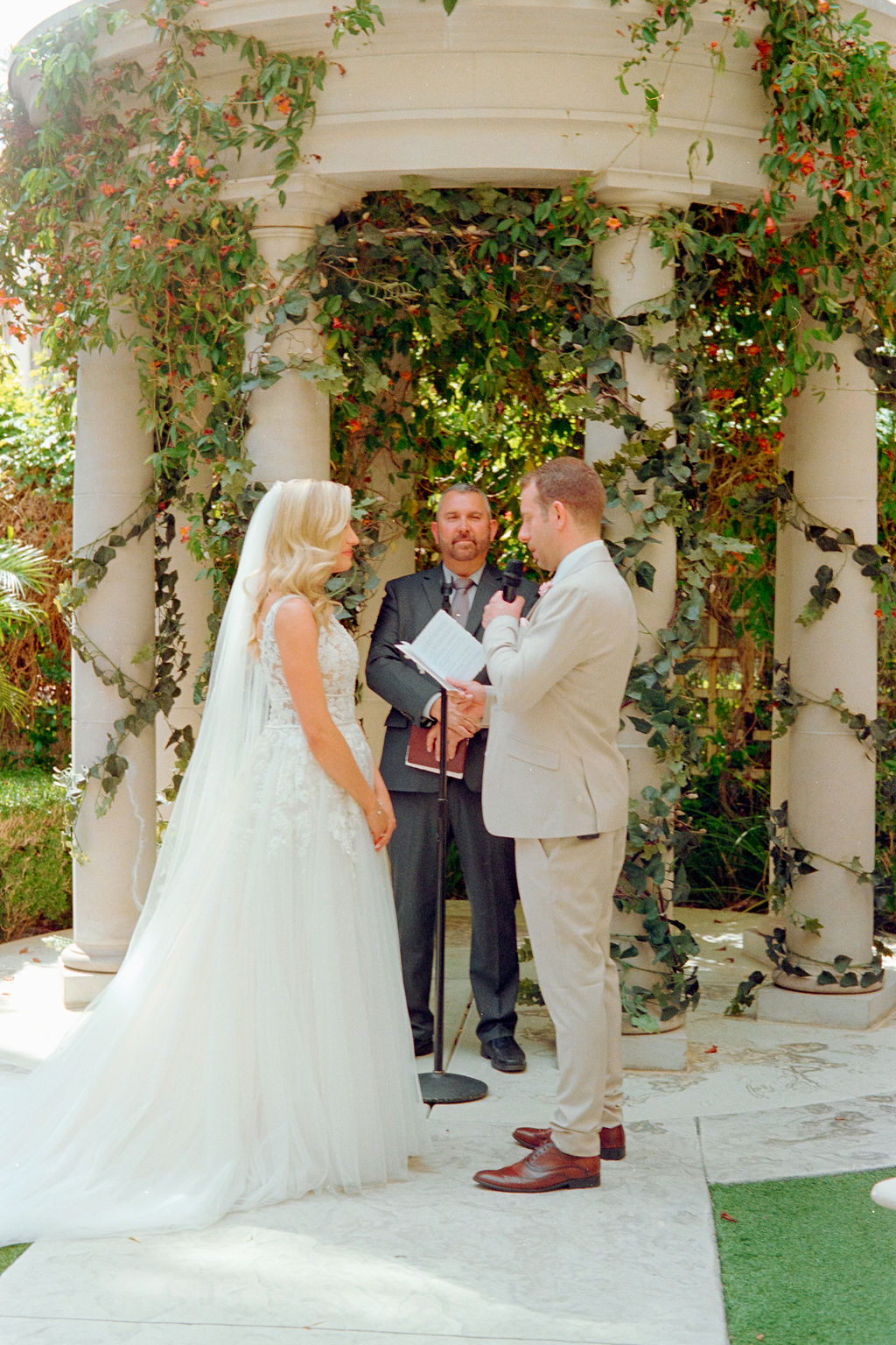 Bride and groom standing under a floral vine-covered gazebo with an officiant during a wedding ceremony.