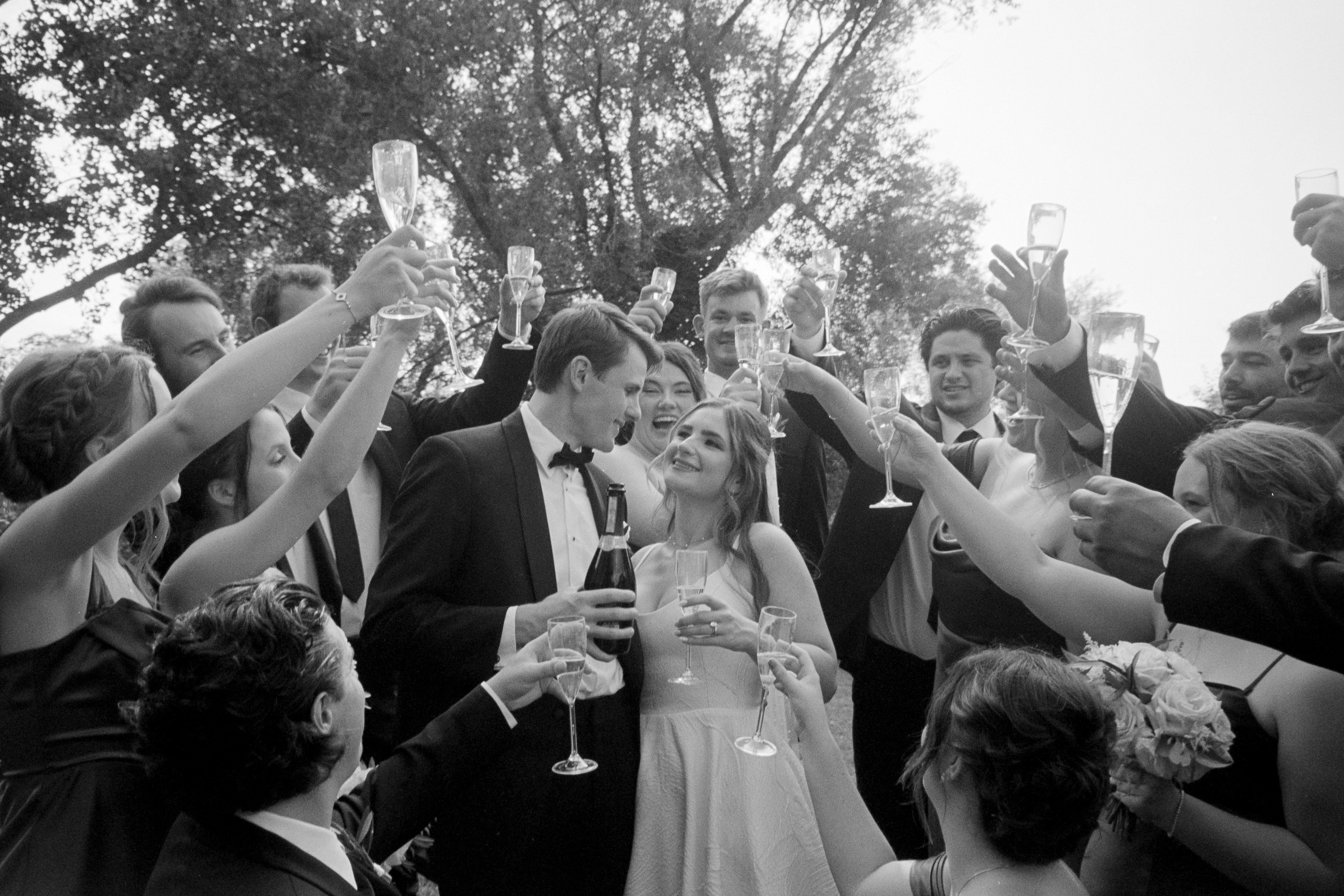 Newlywed couple in wedding attire celebrating and toasting with friends outdoors, holding champagne glasses.