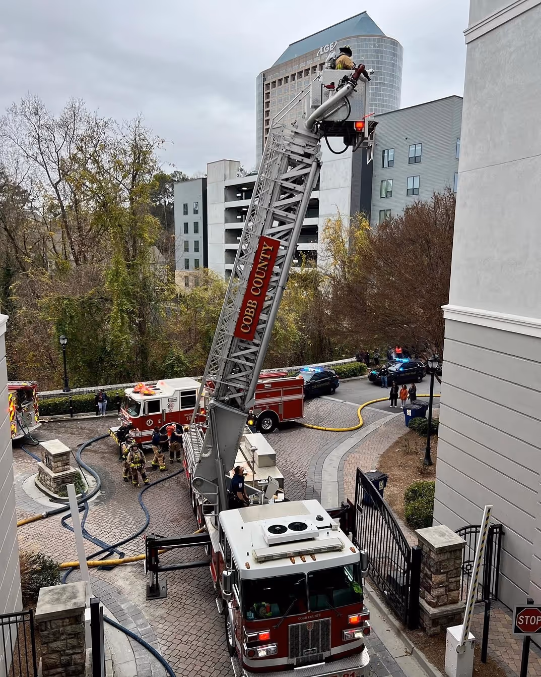 Firefighters using a Cobb County fire truck with an extended ladder at a residential area, with police cars and hoses visible.