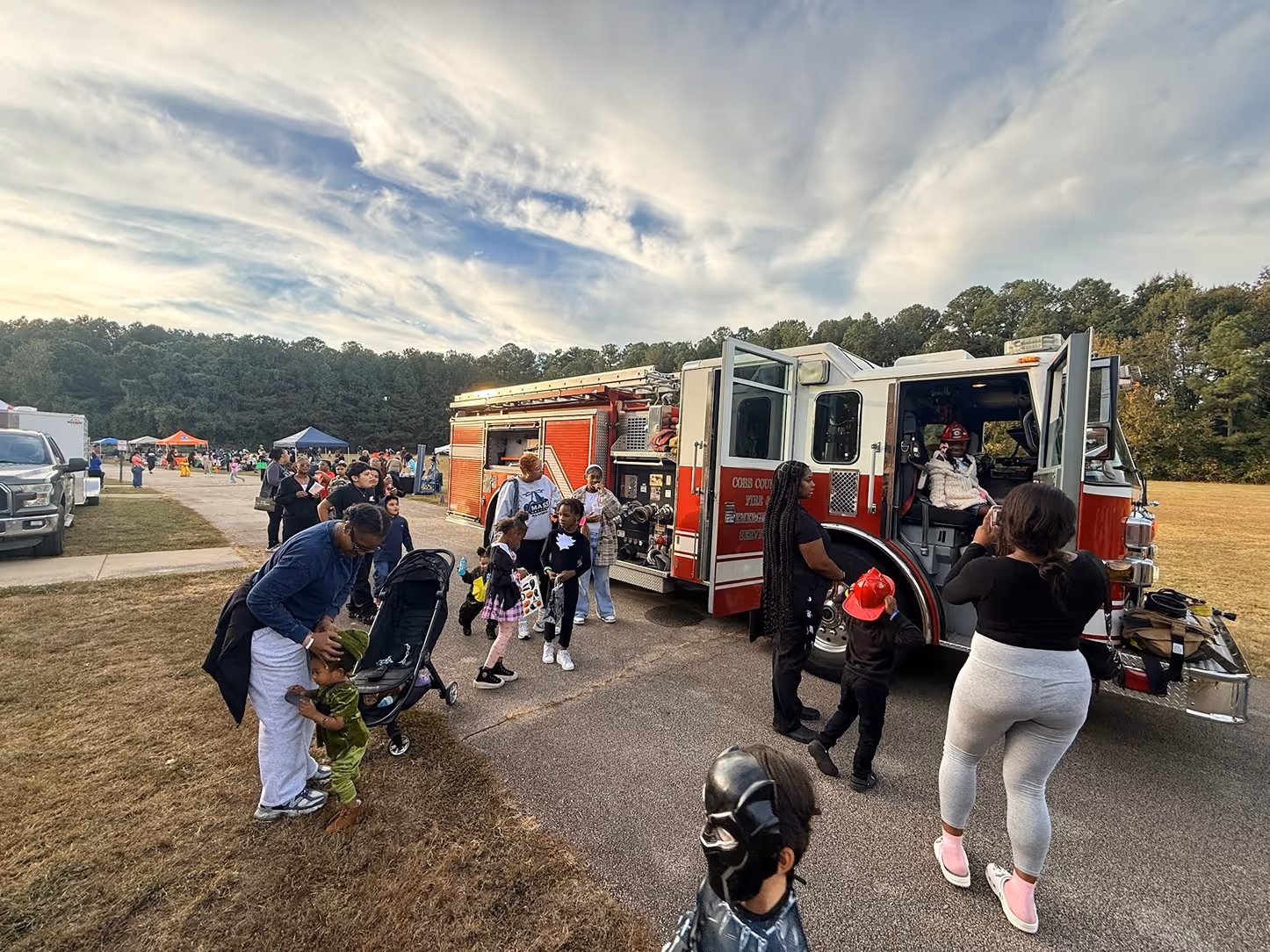 Children and adults gathered around a red fire truck outdoors, with one child wearing a firefighter helmet and another in a costume, under a cloudy sky.