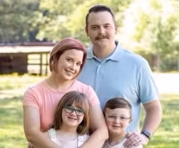 Smiling family of four, including a man, woman, and two children, standing closely together outdoors in a green park.