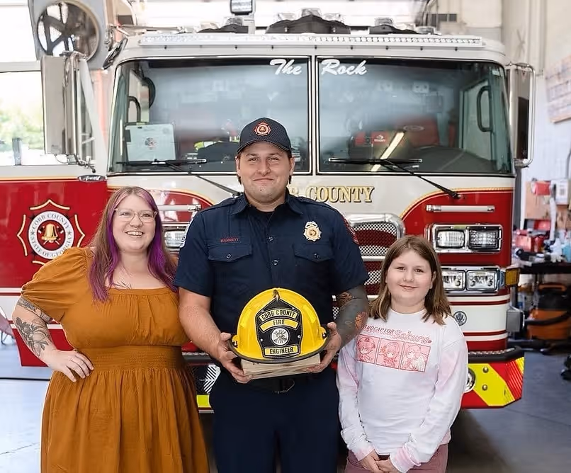 Firefighter in uniform holding a yellow helmet labeled 'Cobb County Fire Engineer' standing between a woman in a mustard dress and a child in a white sweatshirt, in front of a fire truck inside a fire station.