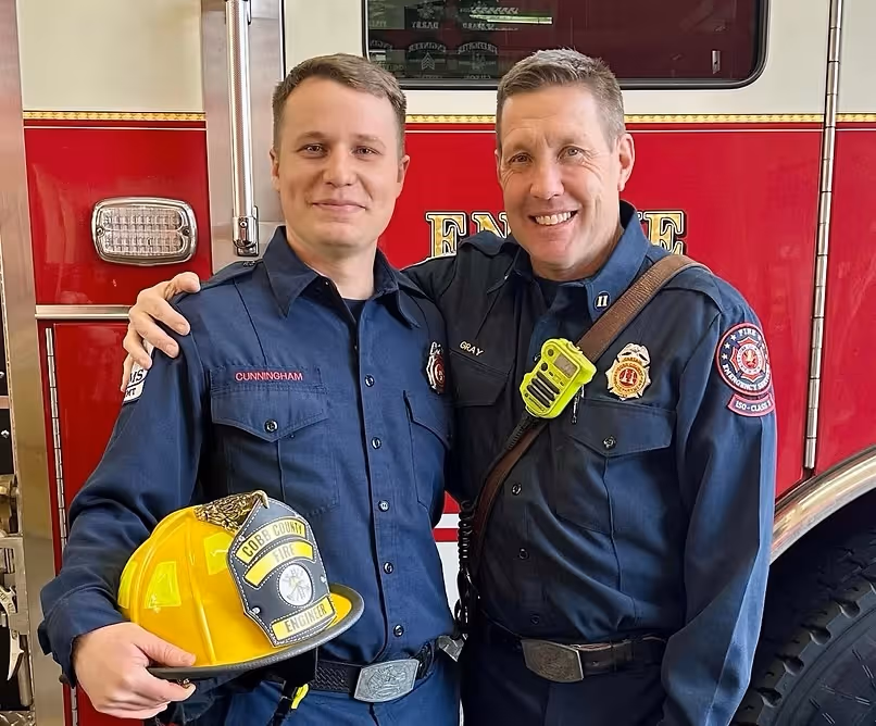 Two firefighters in navy blue uniforms smiling and posing in front of a red fire truck, one holding a yellow fire helmet labeled 'Cobb County Fire Engineer.'
