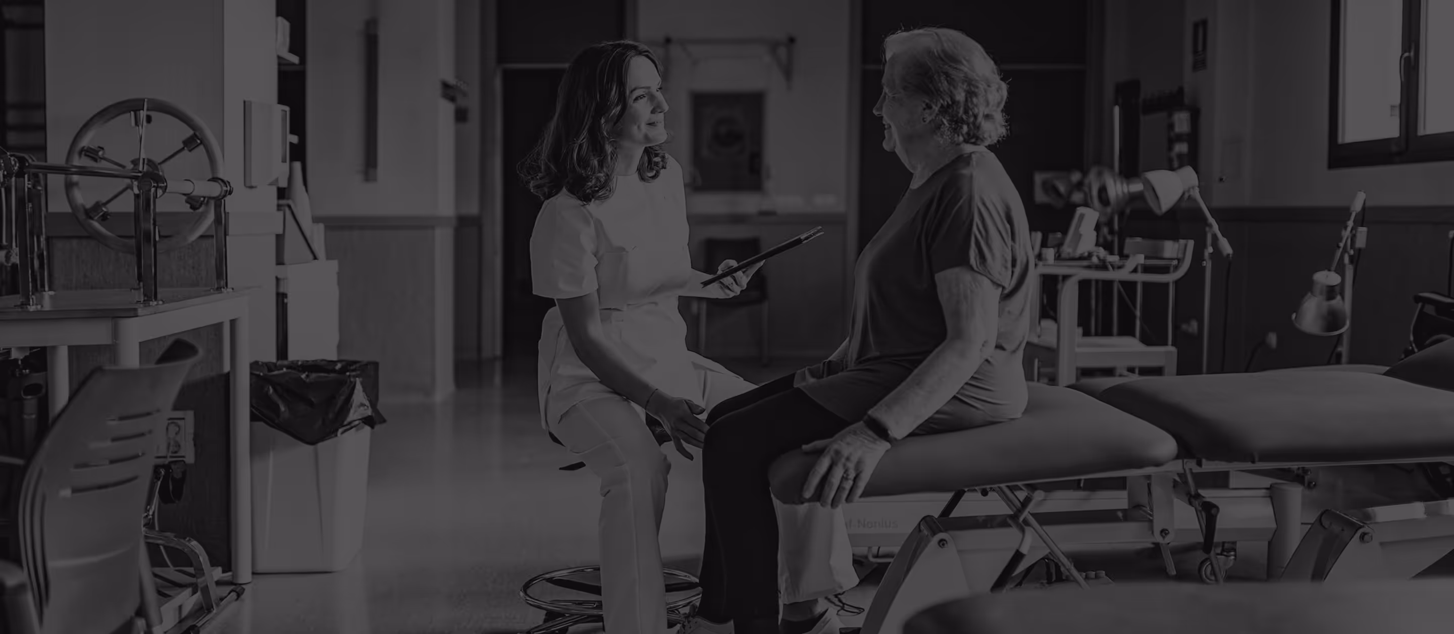 Healthcare professional in white scrubs consulting and smiling at an elderly woman seated on a hospital bed in a clinical room.