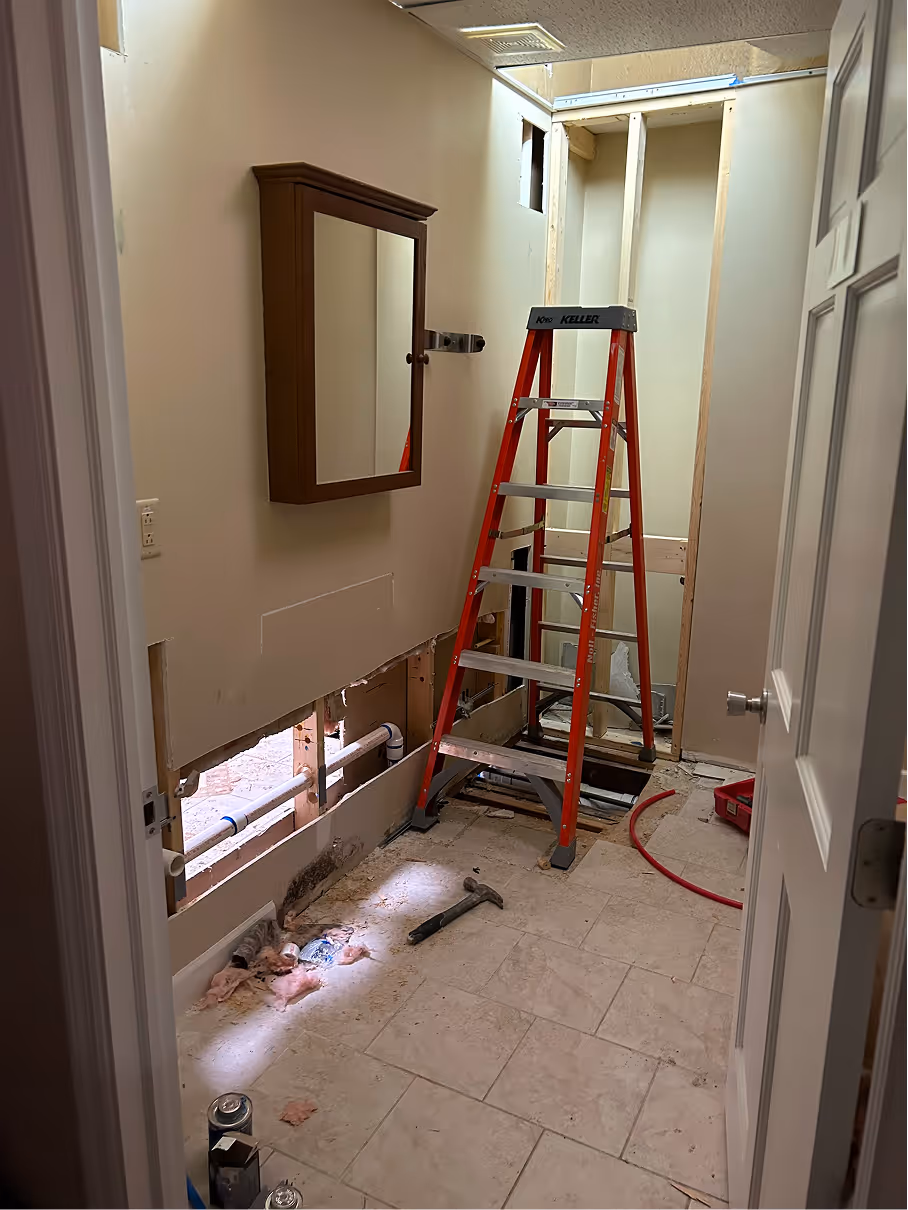 Bathroom renovation in progress showing framed walls, exposed utilities, ladder, and construction tools.