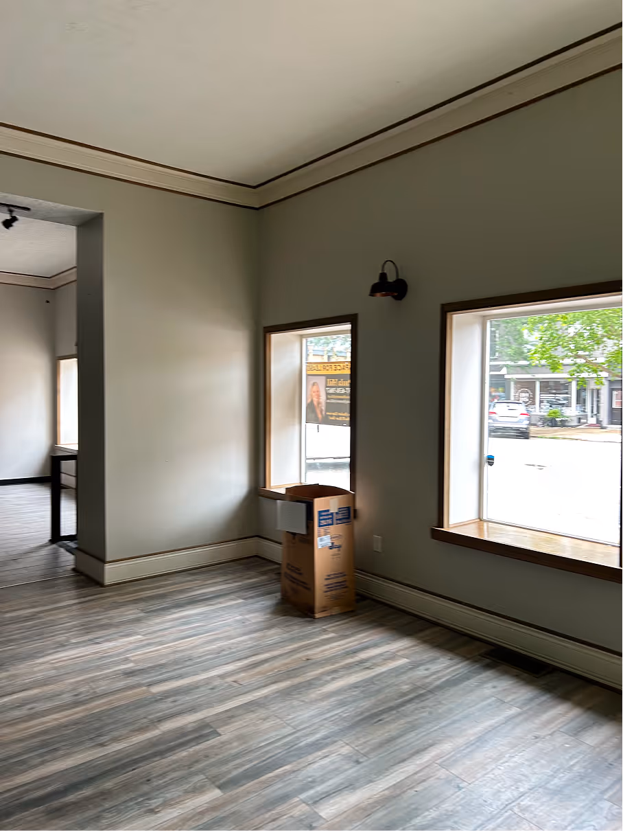 Commercial interior before renovation showing open space, neutral painted walls, large windows, and wood flooring.