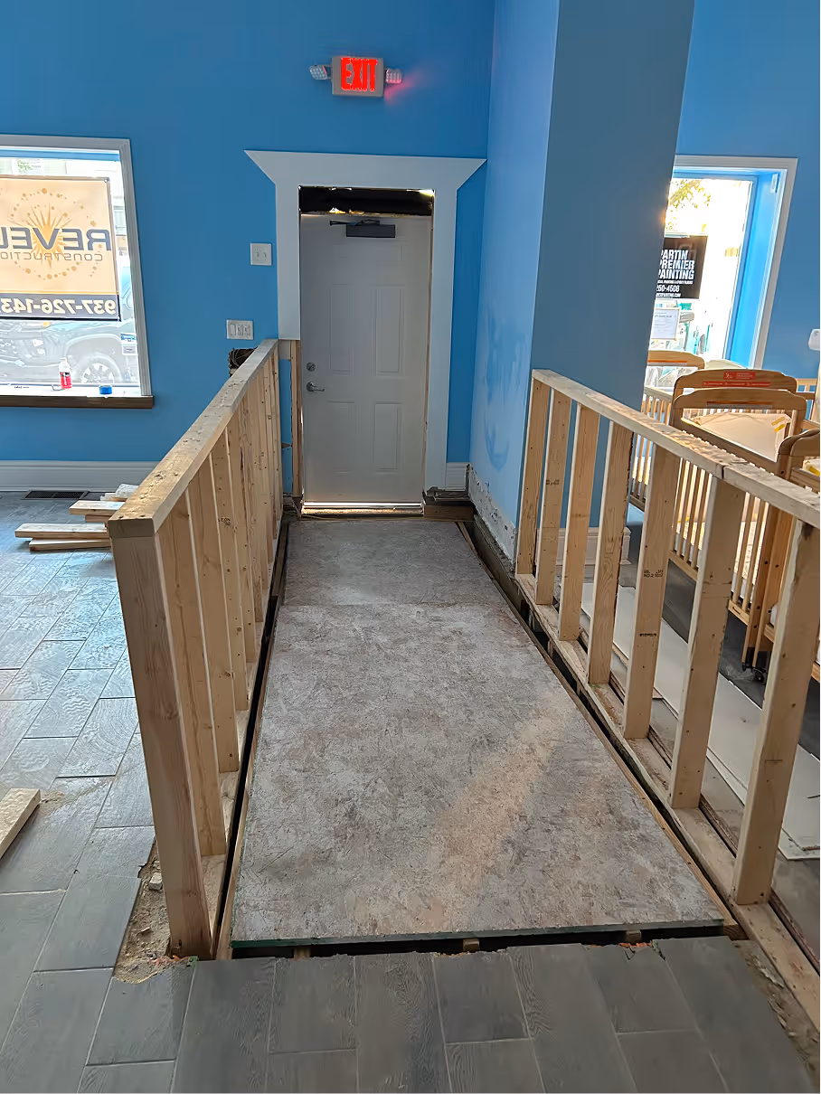 Interior daycare renovation showing a temporary framed accessibility ramp leading to an exit door, with blue painted walls and unfinished flooring.