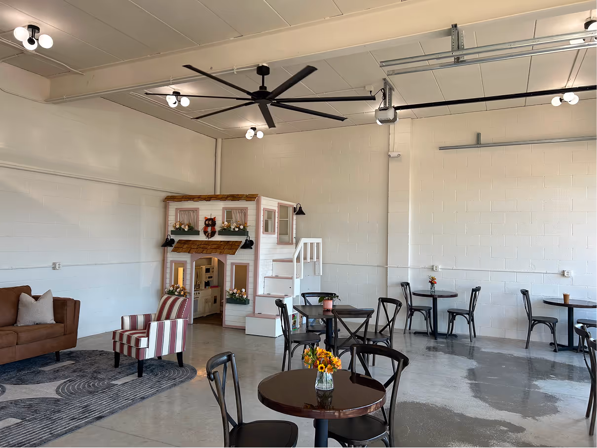 Open commercial space with café tables, decorative playhouse feature, ceiling fans, and polished concrete floor.