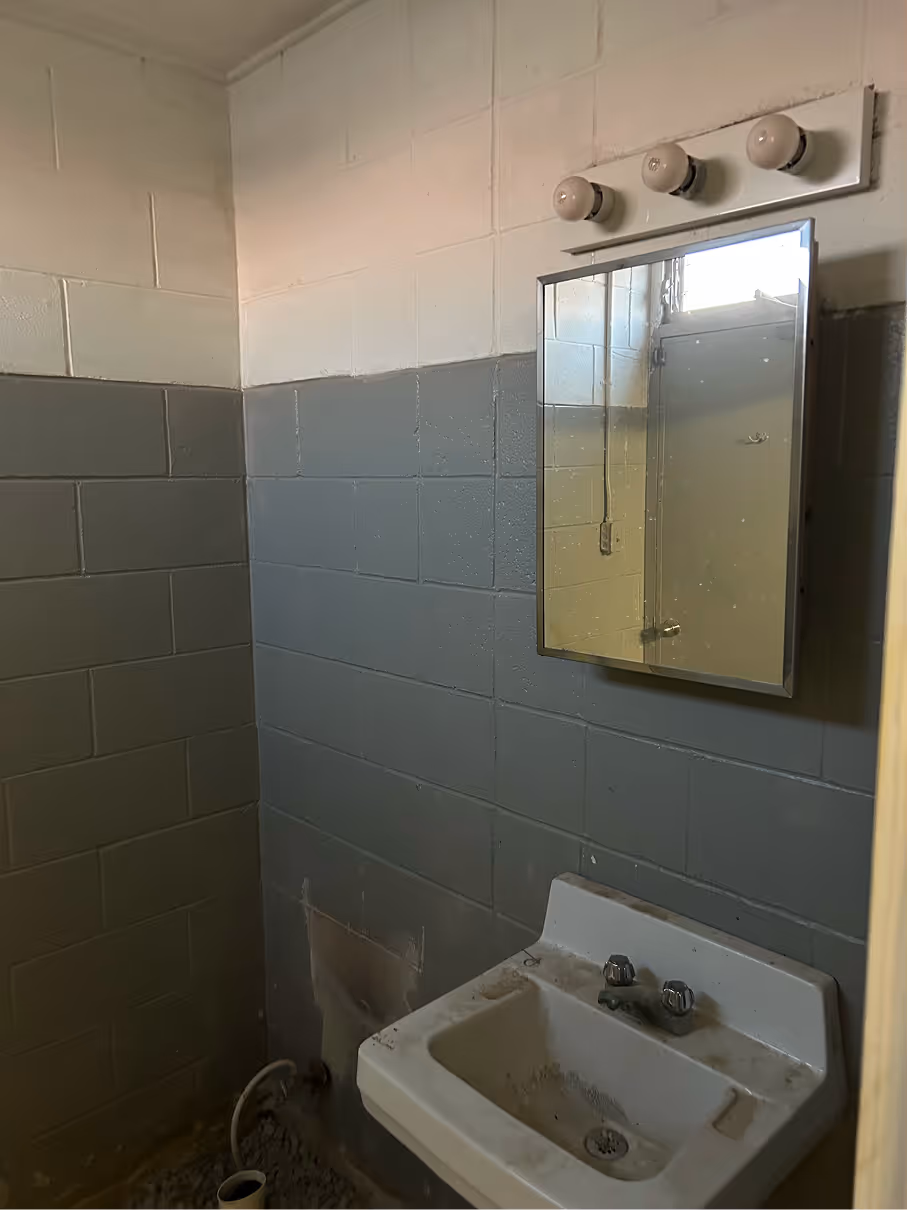 Small restroom before renovation with concrete block walls, wall-mounted sink, mirror, and basic lighting.
