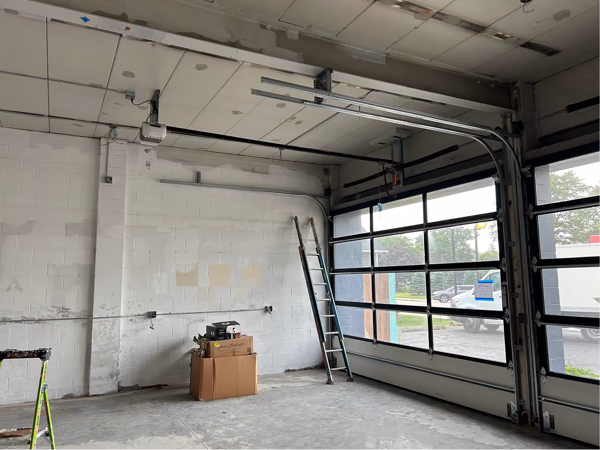 Commercial garage space with glass overhead door, ladder leaning against wall, and unfinished ceiling electrical work.