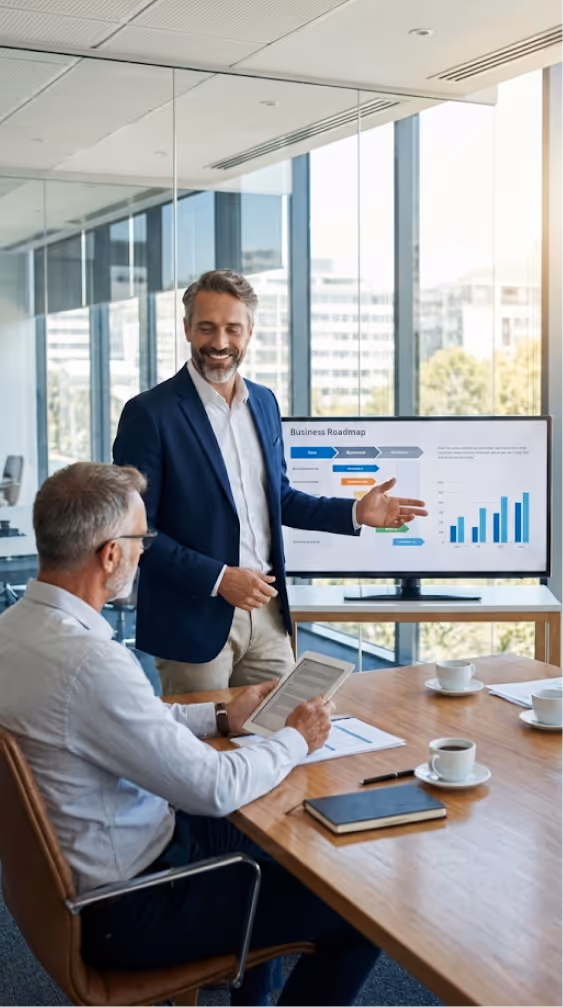 Dwóch mężczyzn na spotkaniu biznesowym w nowoczesnym biurze, jeden prezentuje plan biznesowy na ekranie z wykresem słupkowym, drugi siedzi i trzyma tablet.