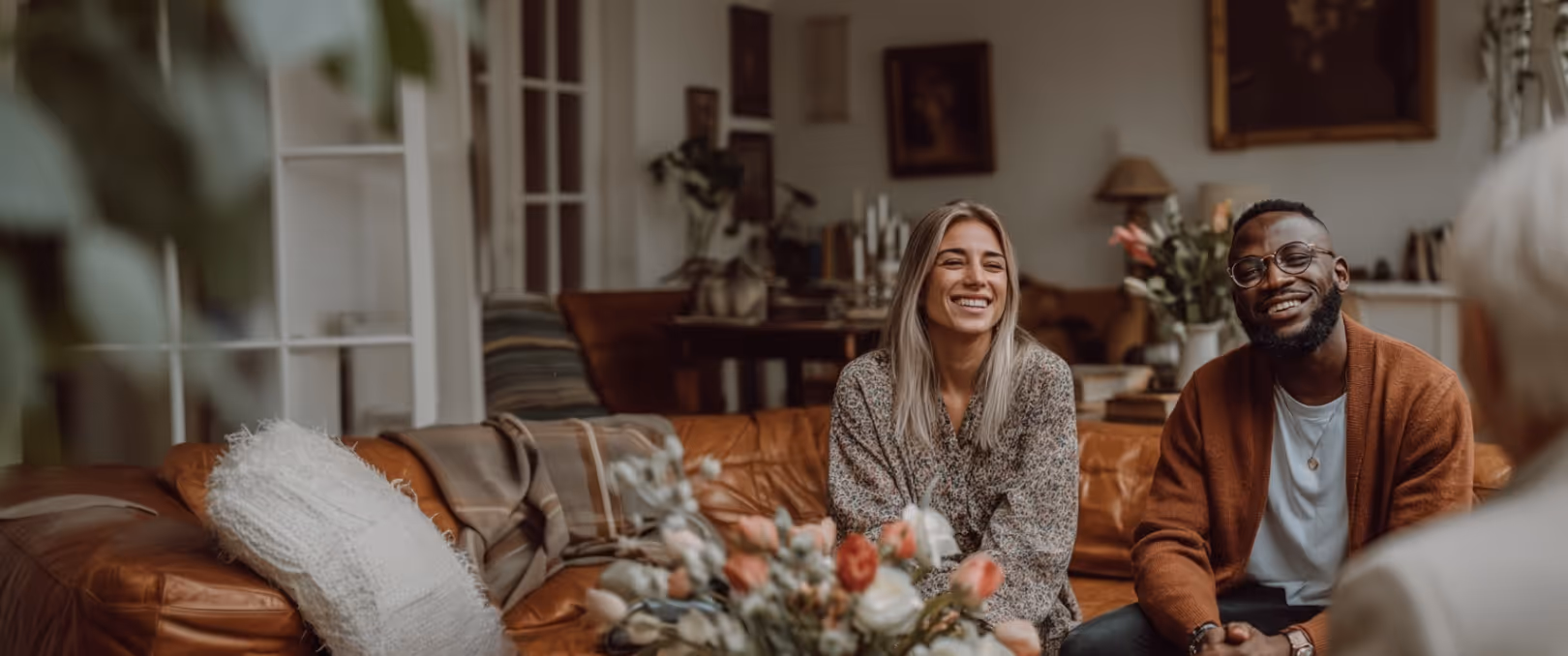 Smiling couple sitting on a leather couch in a cozy living room having a conversation with a person partially visible in the foreground.