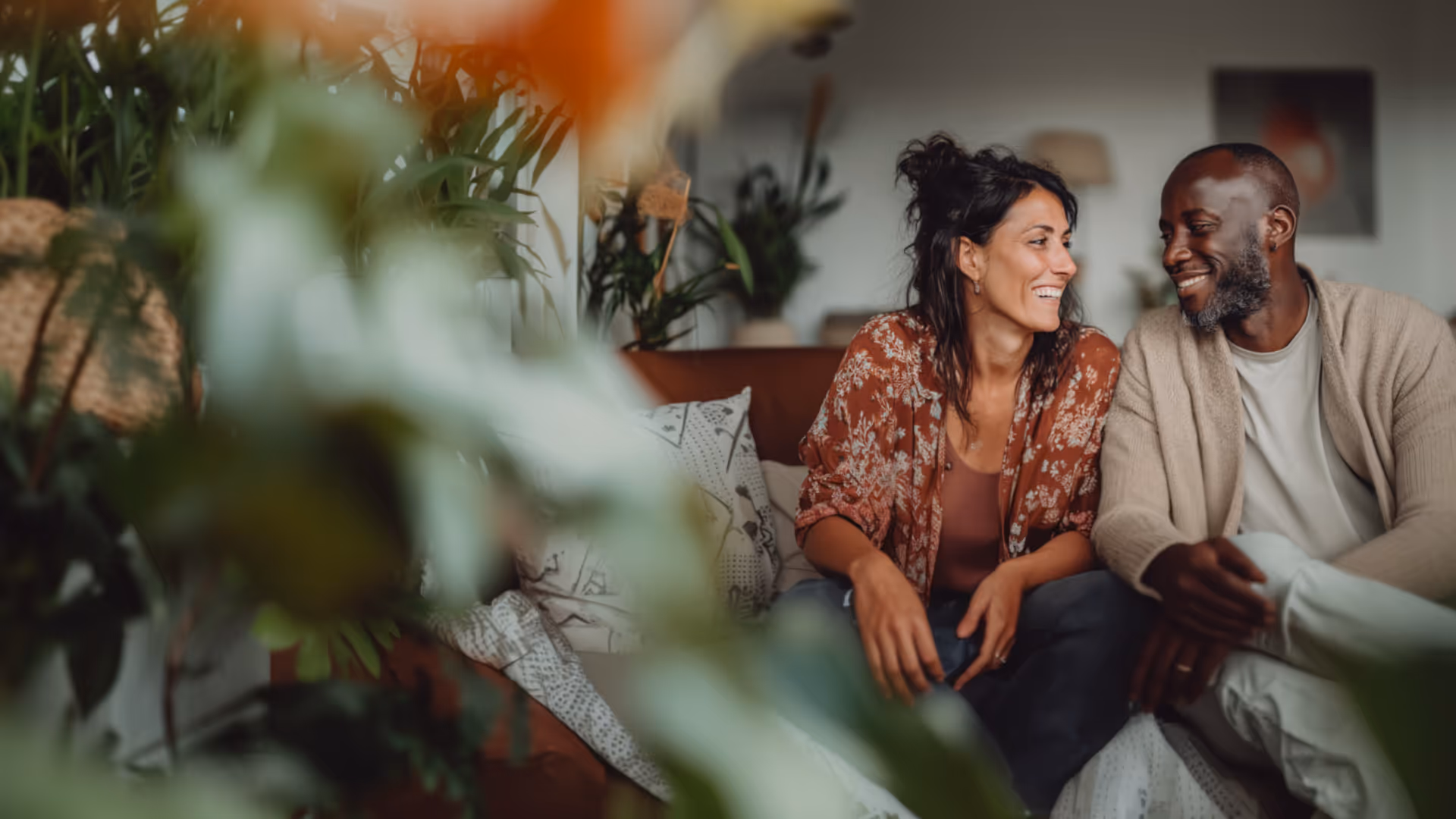 Smiling couple sitting closely on a couch in a cozy, plant-filled living room.