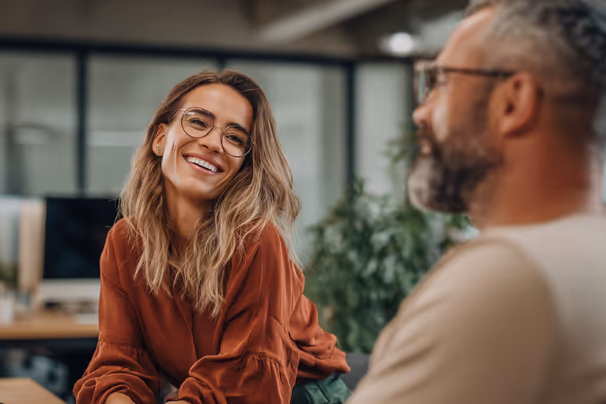 Smiling woman with long hair and glasses engaged in conversation with a man in a casual office setting.