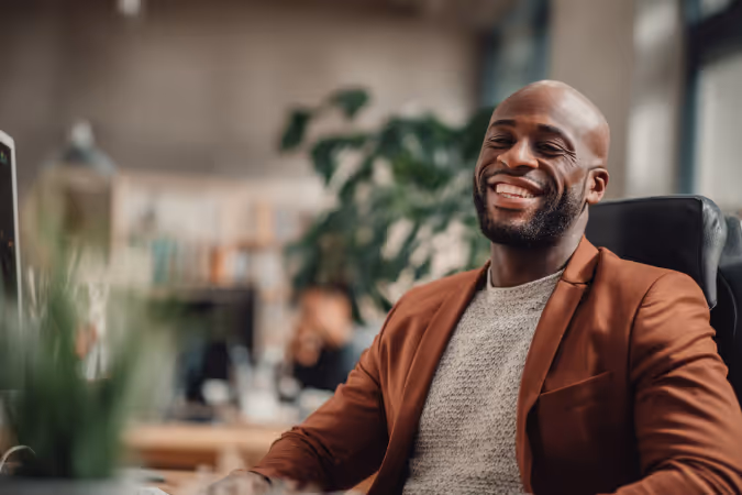 Smiling man wearing a brown jacket sitting in an office with blurred background.