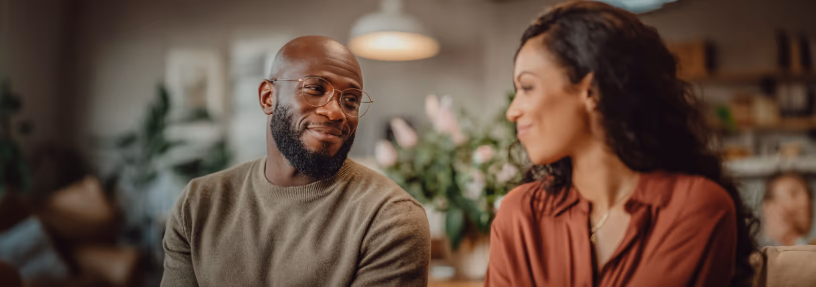 Smiling man with glasses looking affectionately at a woman with curly hair in a cozy indoor setting.