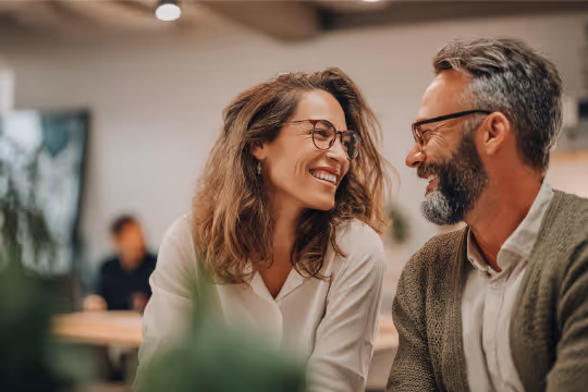 Smiling woman and man wearing glasses looking at each other in a cozy indoor setting.