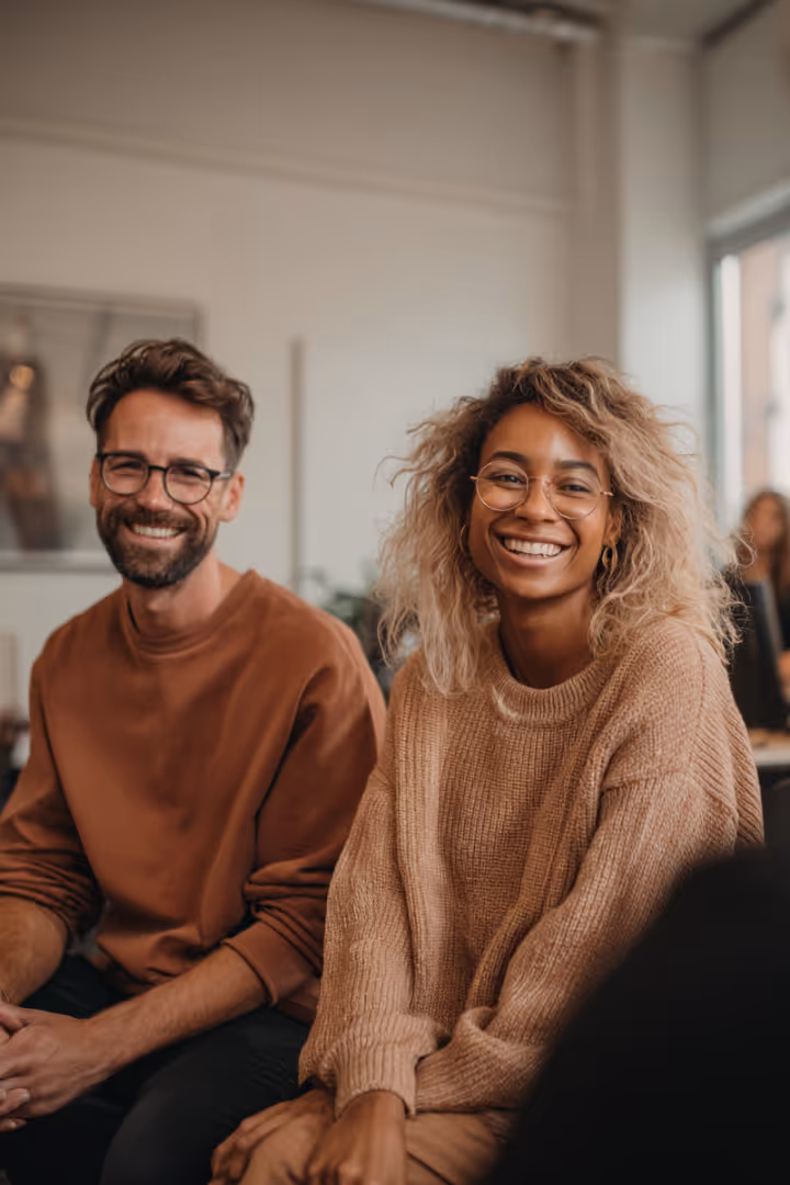 Smiling woman wearing glasses and a sweater sitting at a table with a bearded man in a black shirt in the background.