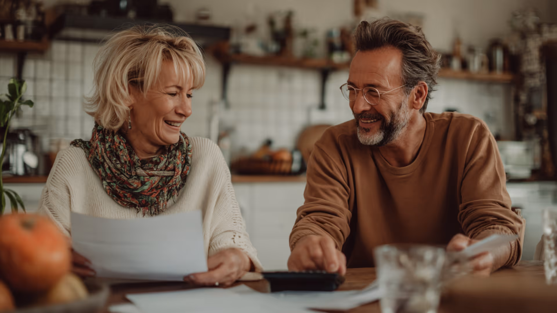 Smiling middle-aged couple sitting at a table in a kitchen, reviewing documents and using a calculator together.