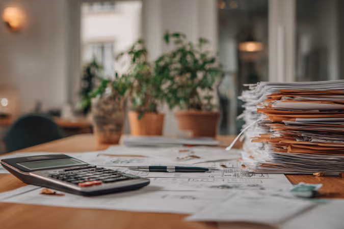 Calculator, pen, paperwork, and a tall stack of files on a wooden desk with blurred potted plants in the background.