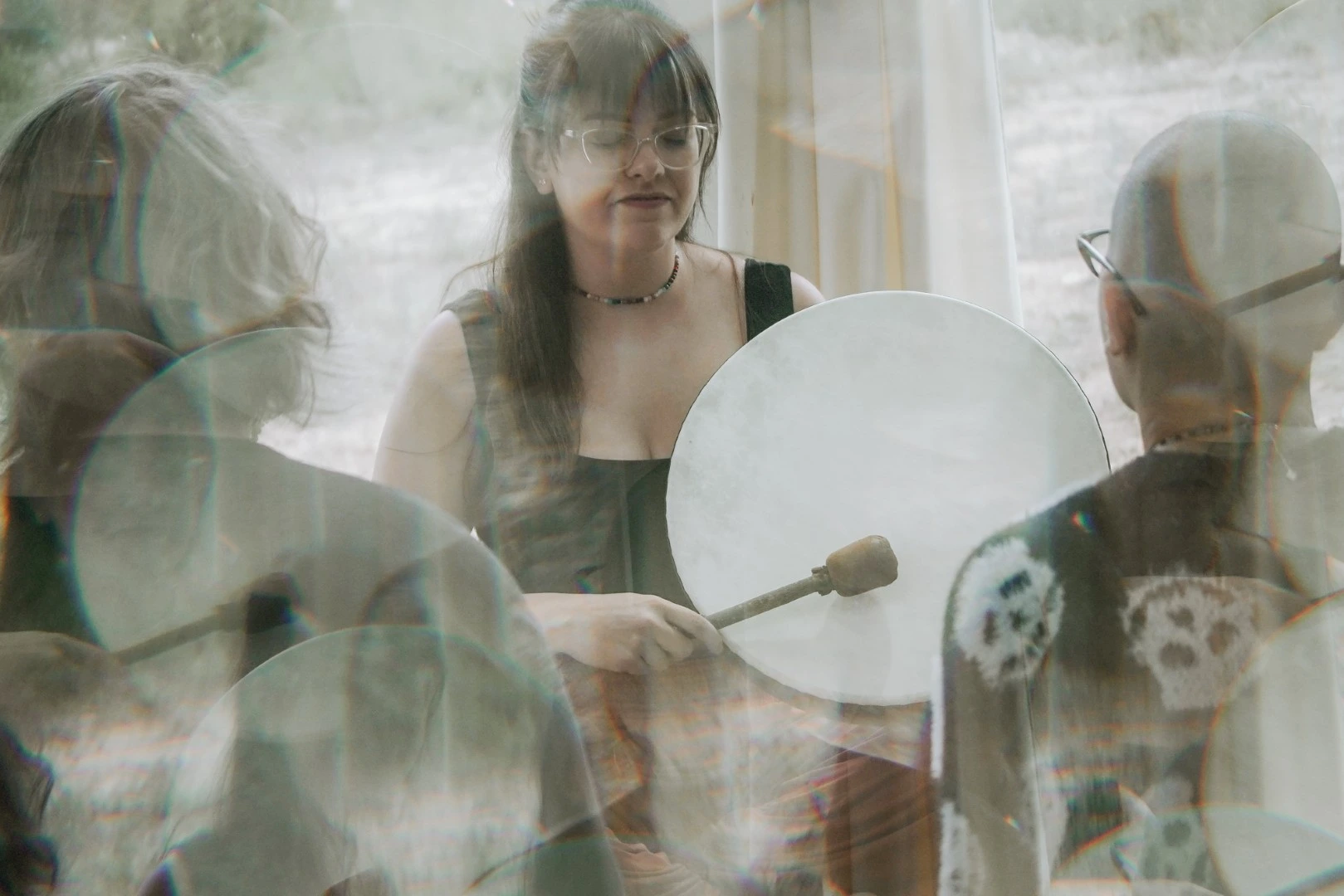 Three people playing handheld frame drums in a softly lit room with a hazy, layered effect.