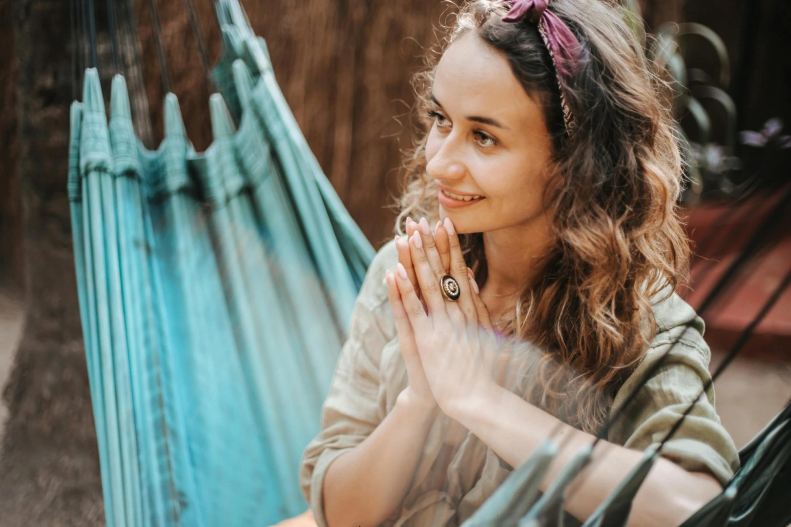 Smiling woman with curly hair and a purple headband sitting in a blue hammock, hands clasped near her face.