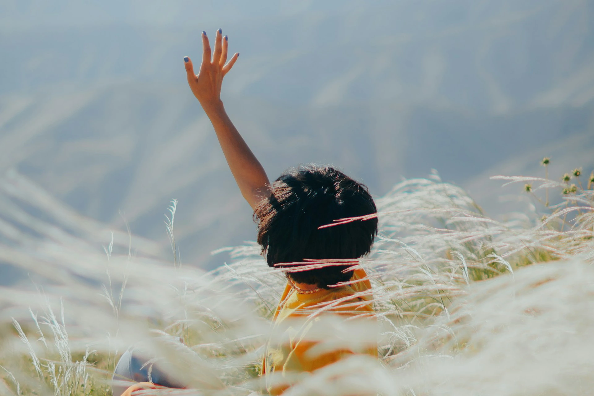 Person with short black hair and blue nail polish raising one hand while sitting in a field of tall grasses with mountains in the background.