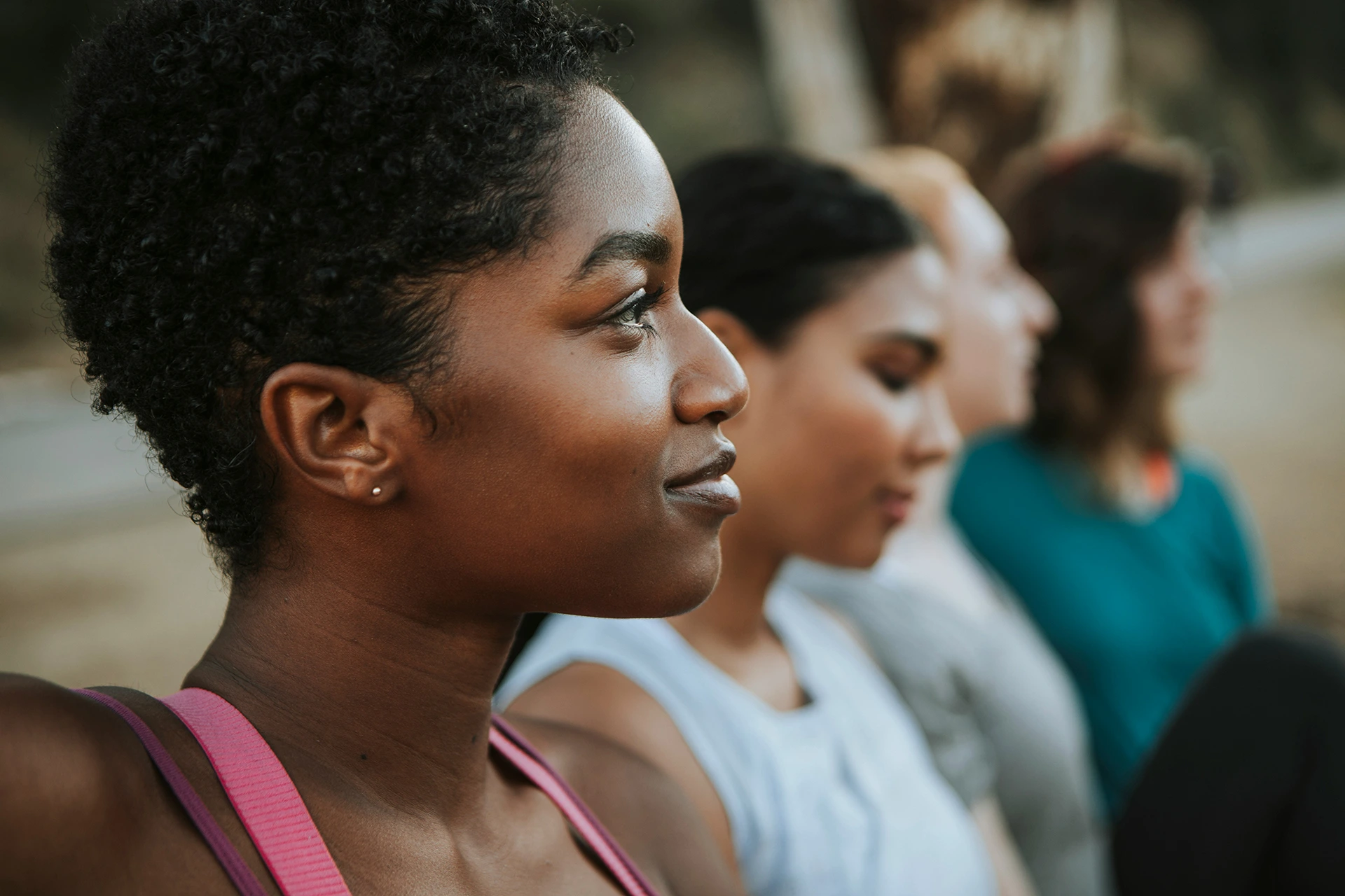 Close-up of a smiling young Black woman with short curly hair in profile, with three blurred people sitting in a row behind her.