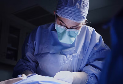 Surgeon wearing surgical mask and cap intensely performing an operation under bright light in a dimly lit room.
