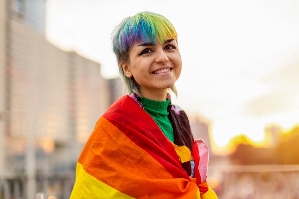 A woman with short coloured hair wears a rainbow flag around her shoulders with a sunny background. 