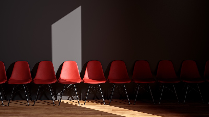 a dark grey wall with wooden floor mostly in shadow. A sliver of sun shines through onto a row of bright red chairs. 
