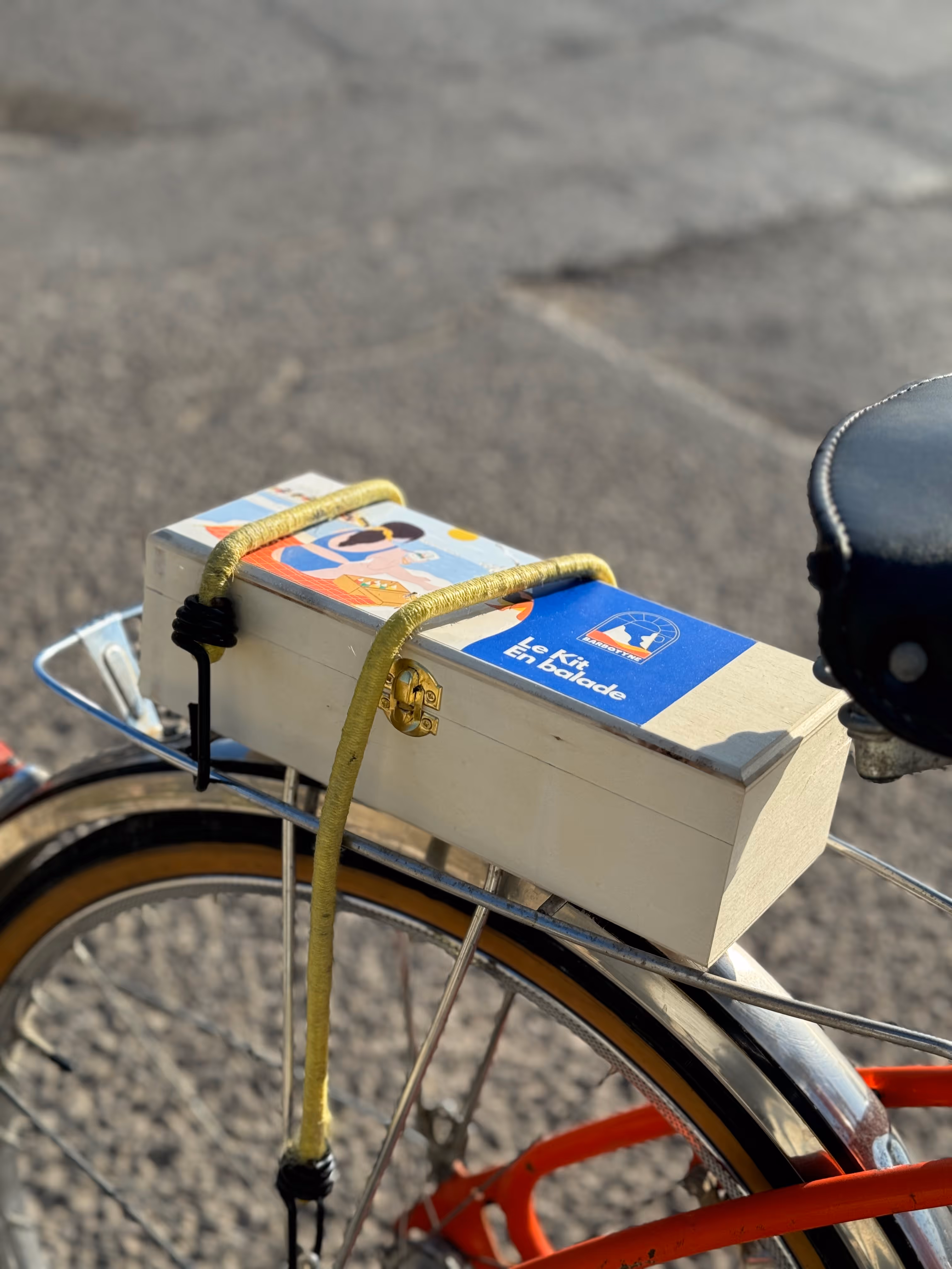Boîte métallique blanche avec un autocollant bleu « Le Kit En balade » et une poignée recouverte de corde jaune.
