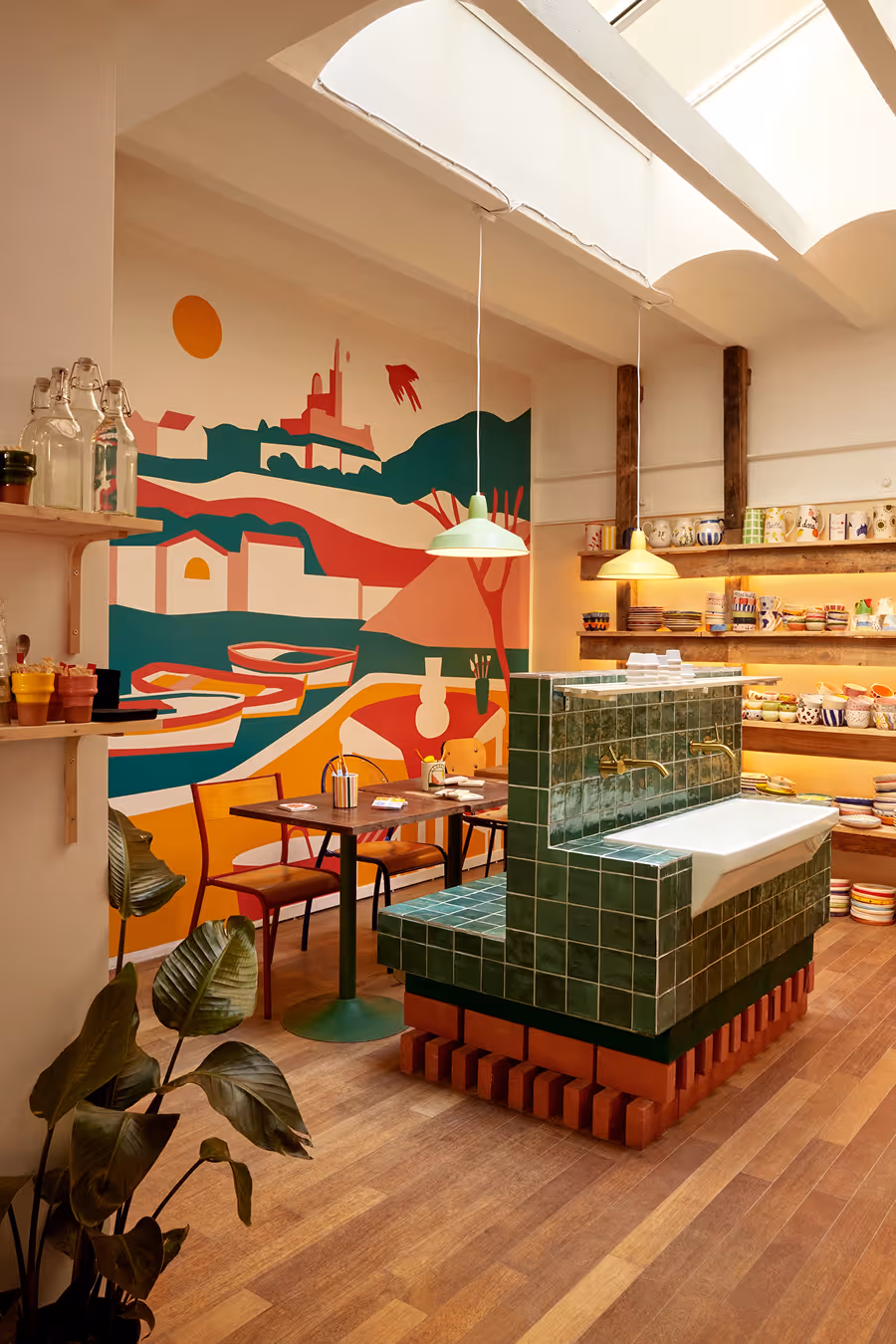 Intérieur d'un café avec murs décorés d'une fresque colorée, tables en bois, et un grand lavabo carrelé vert au centre.
