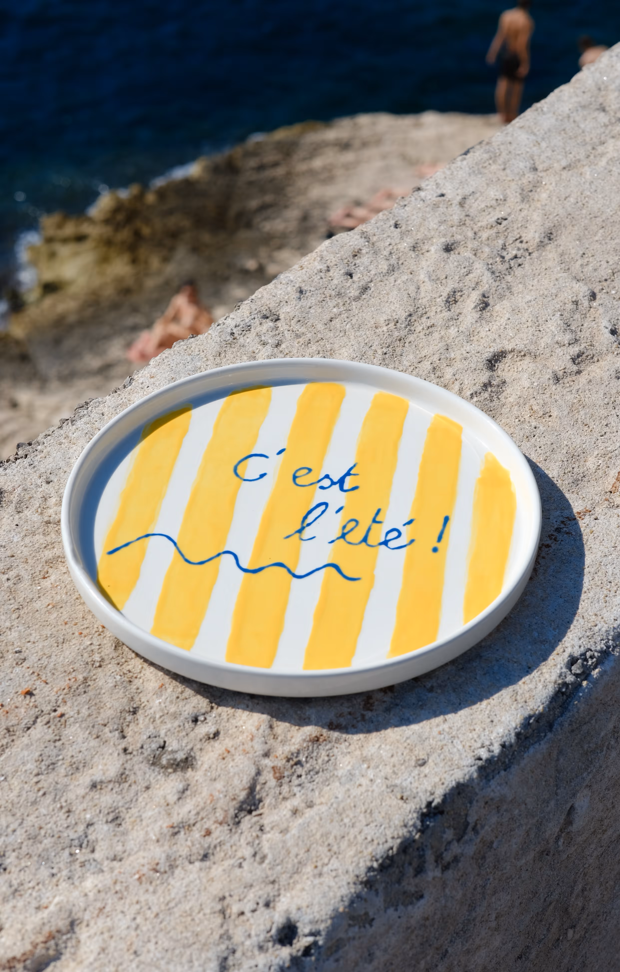 Une assiette ronde avec des rayures jaunes et blanches et le texte « C'est l'été ! » en bleu, posée sur un mur en pierre au bord de la mer.