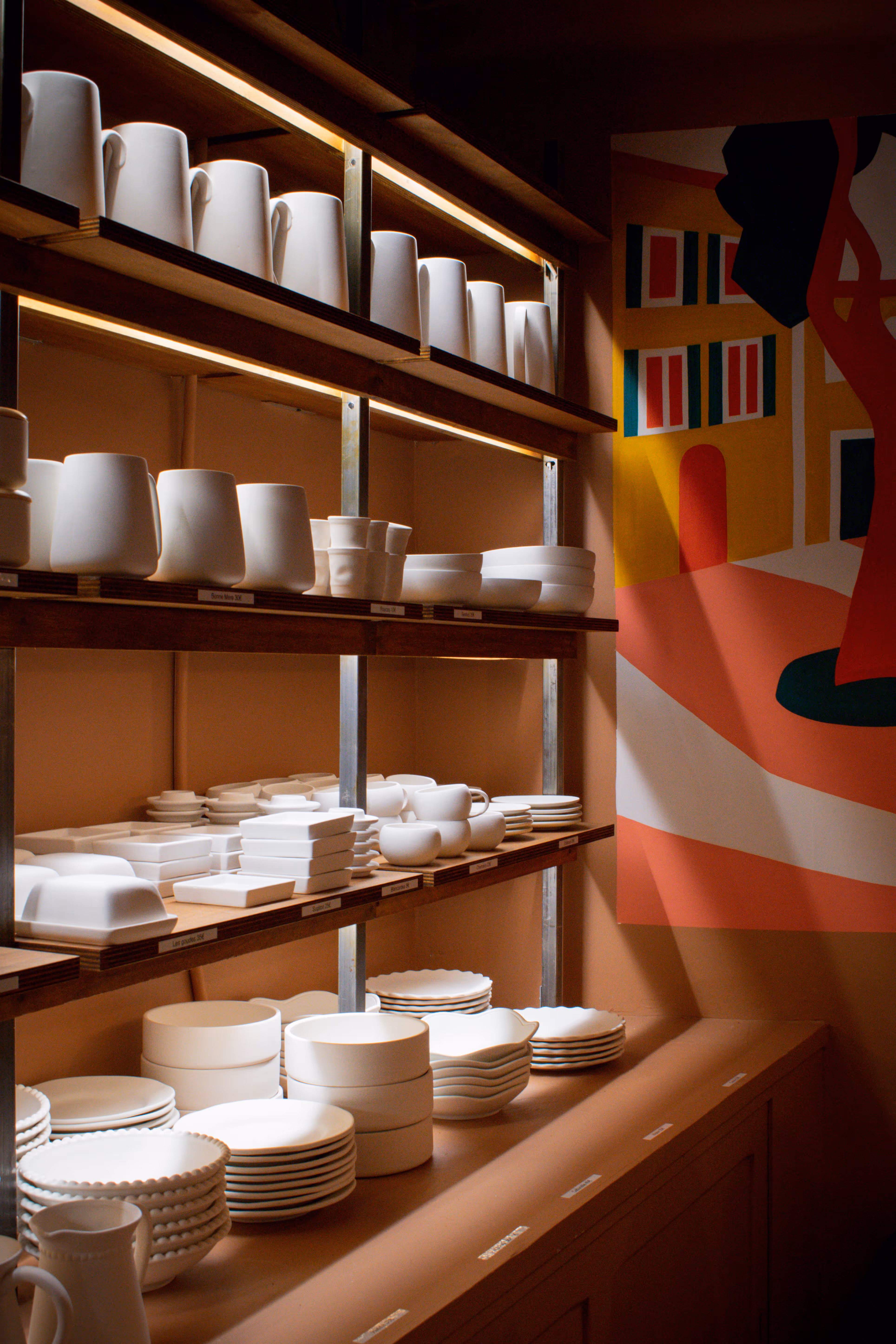 Étagères en bois avec vaisselle blanche mate empilée et disposée dans une pièce aux murs bruns avec une peinture murale colorée à droite.