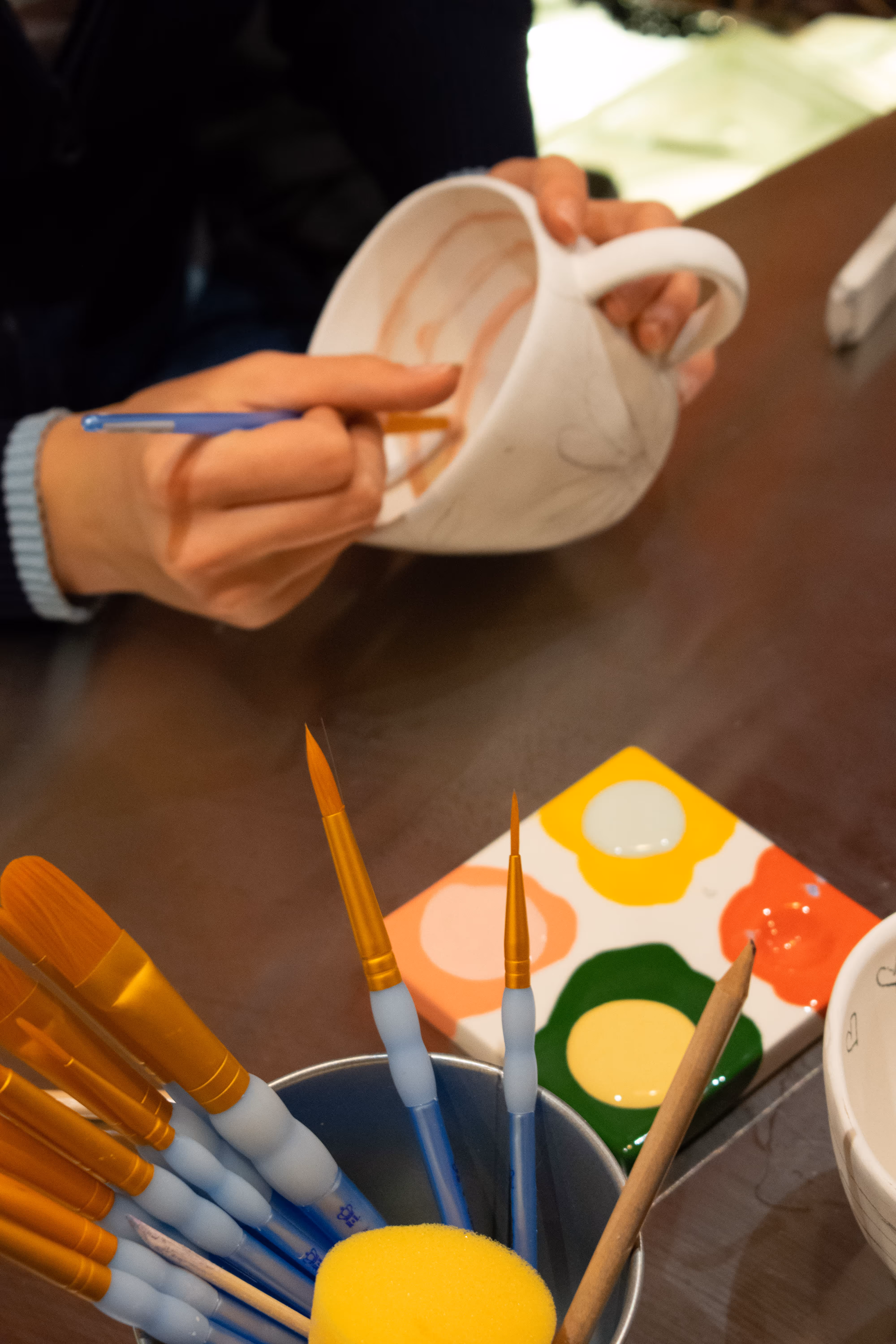 Main tenant un pinceau et peignant l'intérieur d'une tasse blanche, avec des pinceaux et une palette de peinture colorée sur la table.