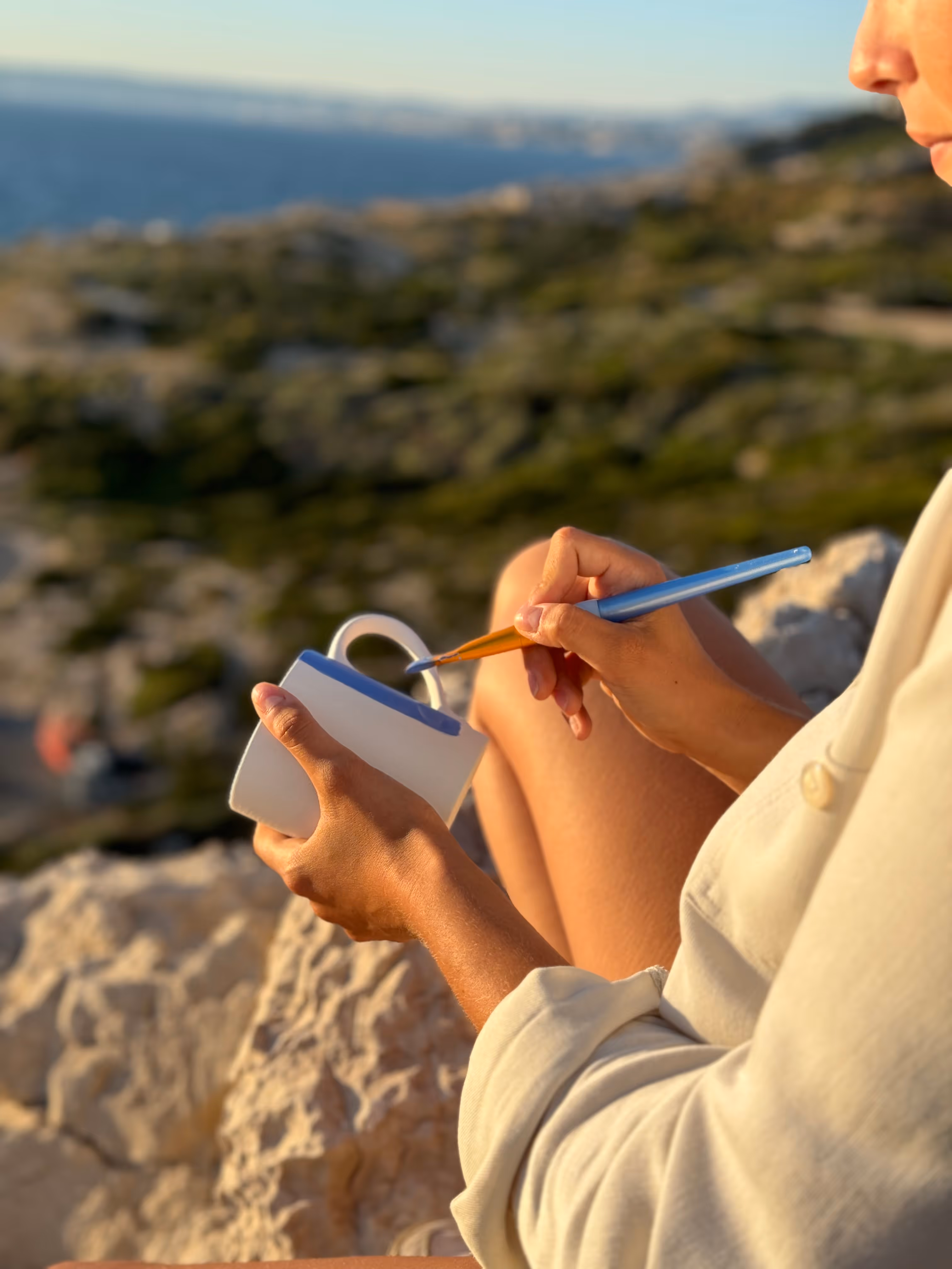 Person peignant le bord d'une tasse blanche en plein air avec une mer et des rochers en arrière-plan.