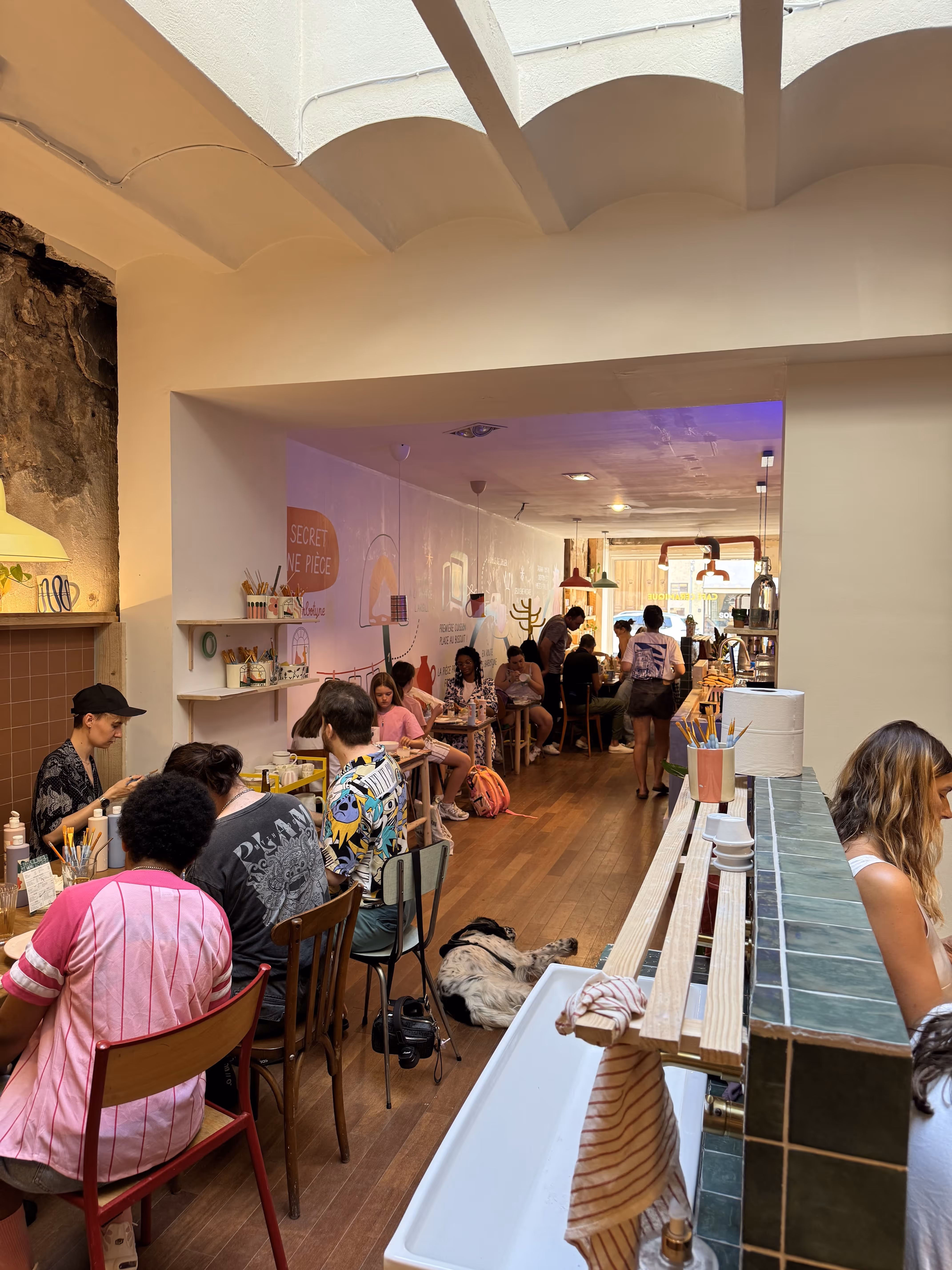 Un café animé avec des personnes assises à une longue table en bois, peignant, avec un chien couché sur le sol en bois sous la table.