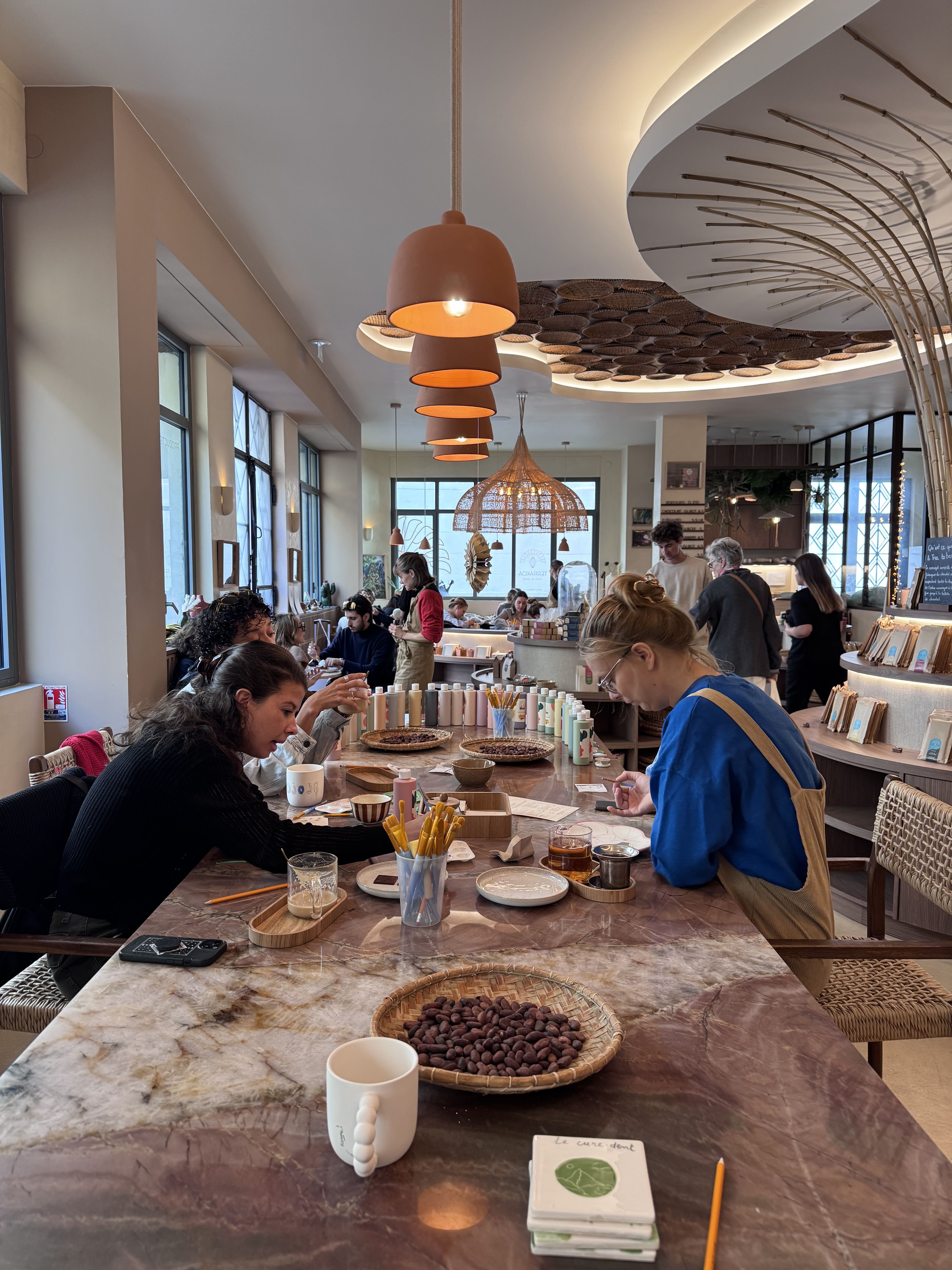 Des personnes assises autour d'une grande table en marbre dans un café lumineux, décoré avec des suspensions en terre cuite et des paniers décoratifs au plafond.