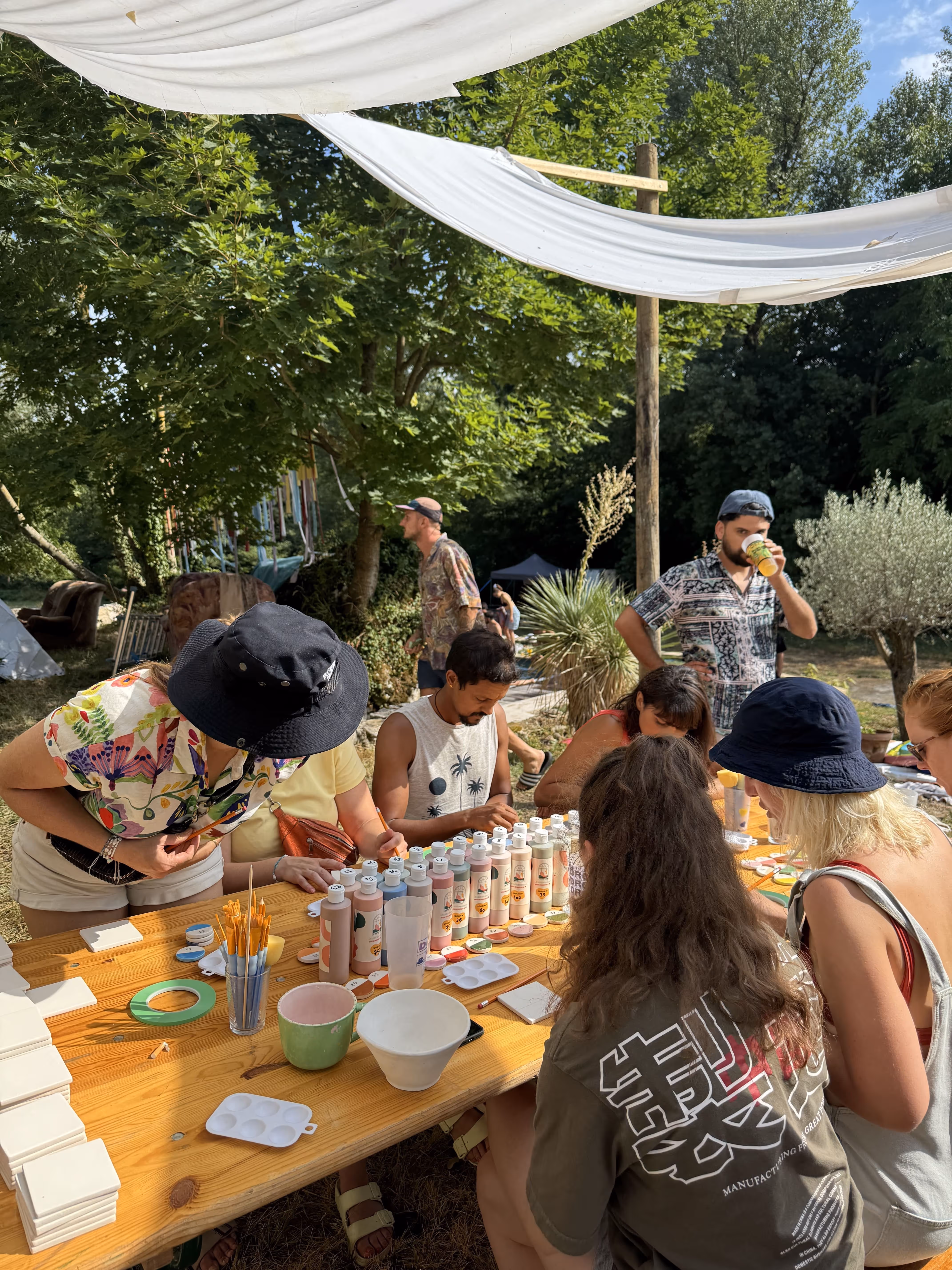 Groupe de personnes assises autour d'une table en bois en plein air, participant à une activité de peinture avec des bouteilles de peinture et des pinceaux sous un auvent blanc.