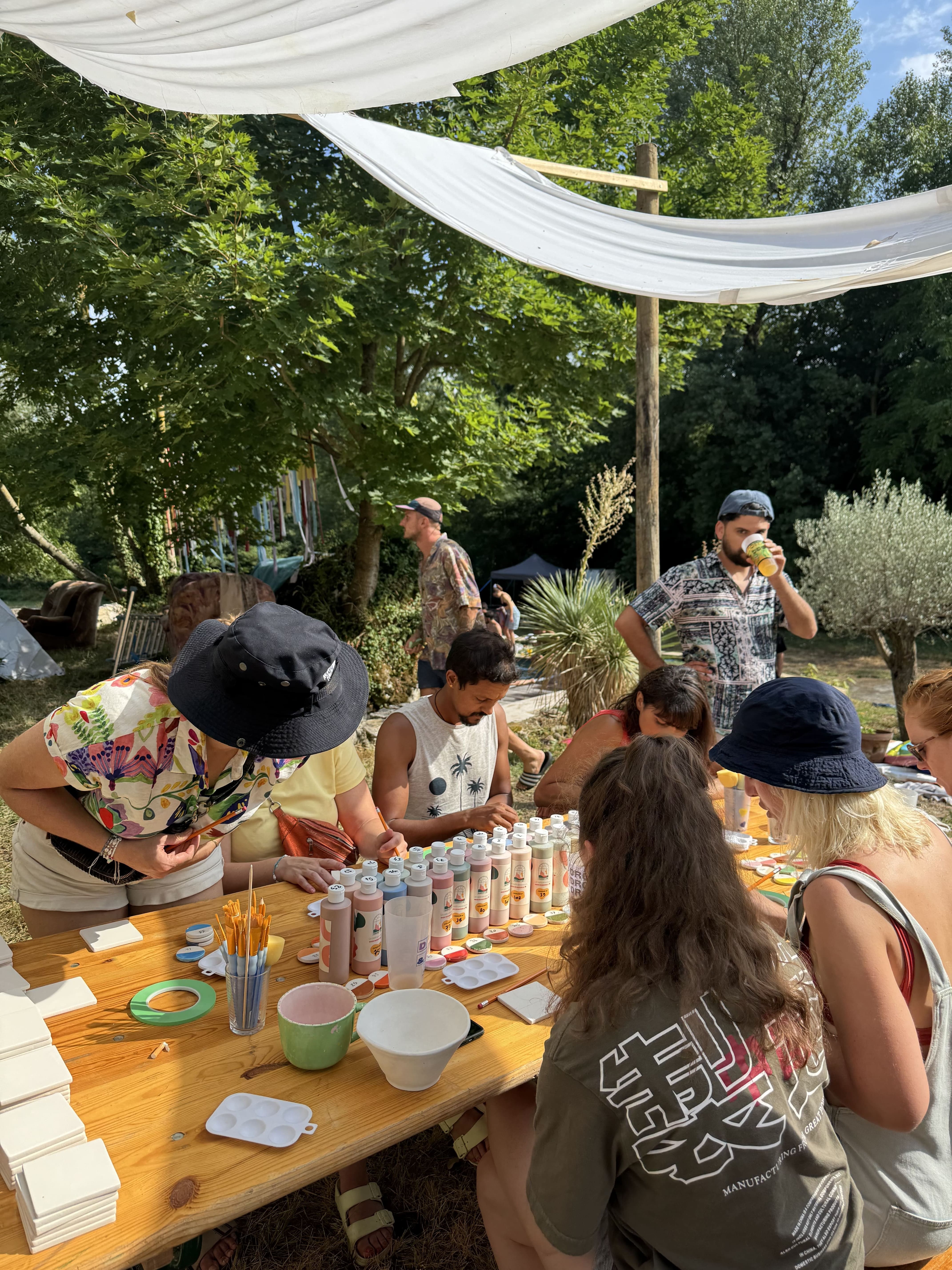 Groupe de personnes assises autour d'une table en bois en plein air, participant à une activité de peinture avec des bouteilles de peinture et des pinceaux sous un auvent blanc.