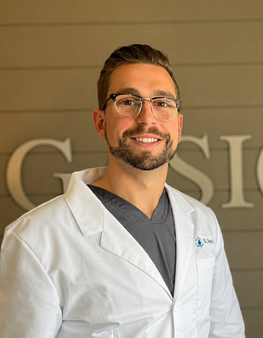 Dr. Max Gosselin, a dentist at Gasior Family Dental, wearing glasses and a white lab coat over gray scrubs, standing indoors against a neutral wall with partial lettering visible in the background.